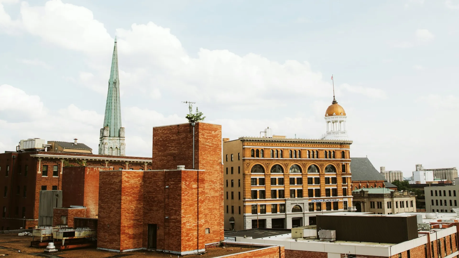 Cityscape featuring historic brick buildings, a church steeple, and a domed tower under a partly cloudy sky.