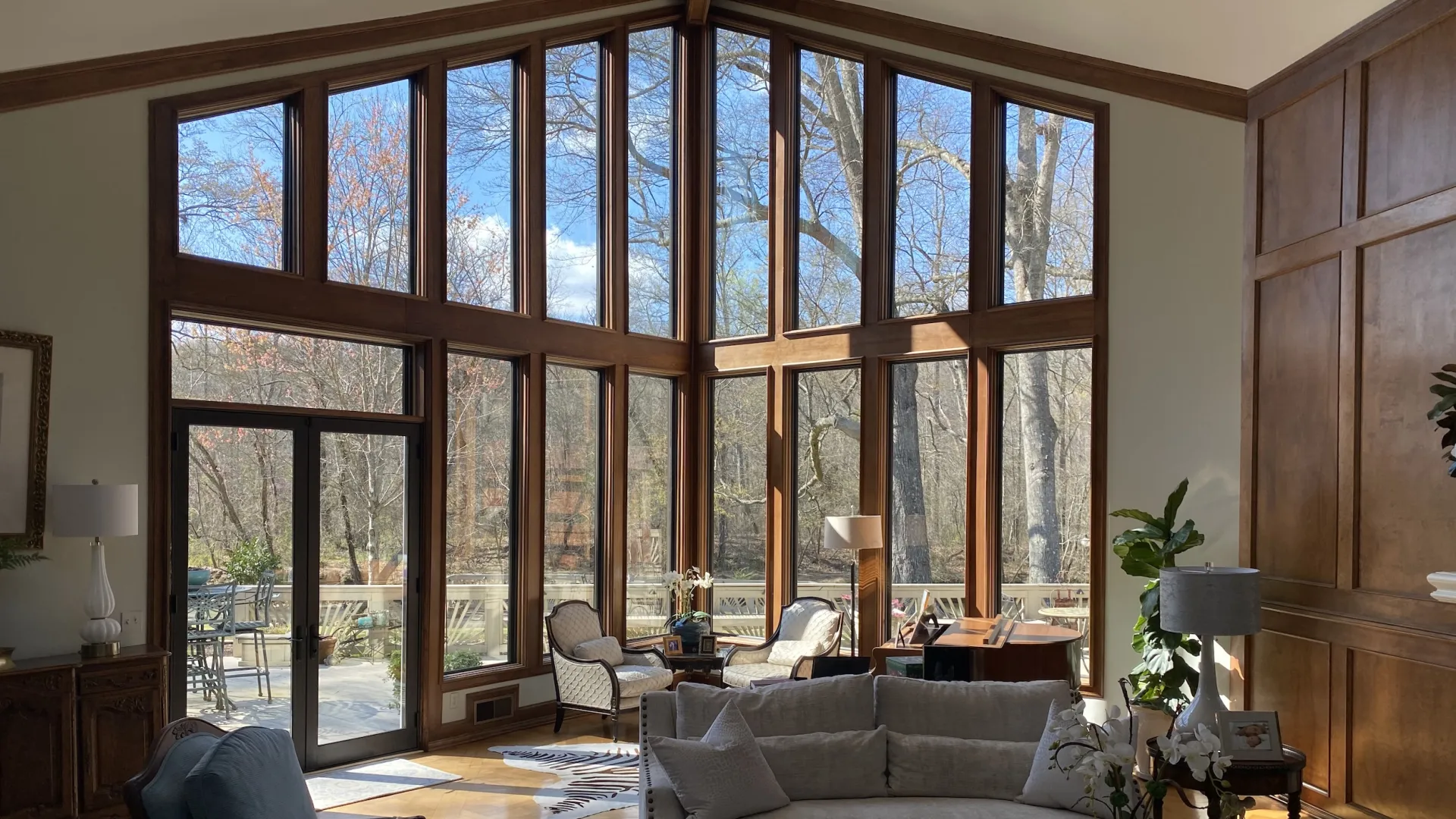 Spacious living room with tall wood-framed windows overlooking a wooded outdoor patio area, natural light floods interior