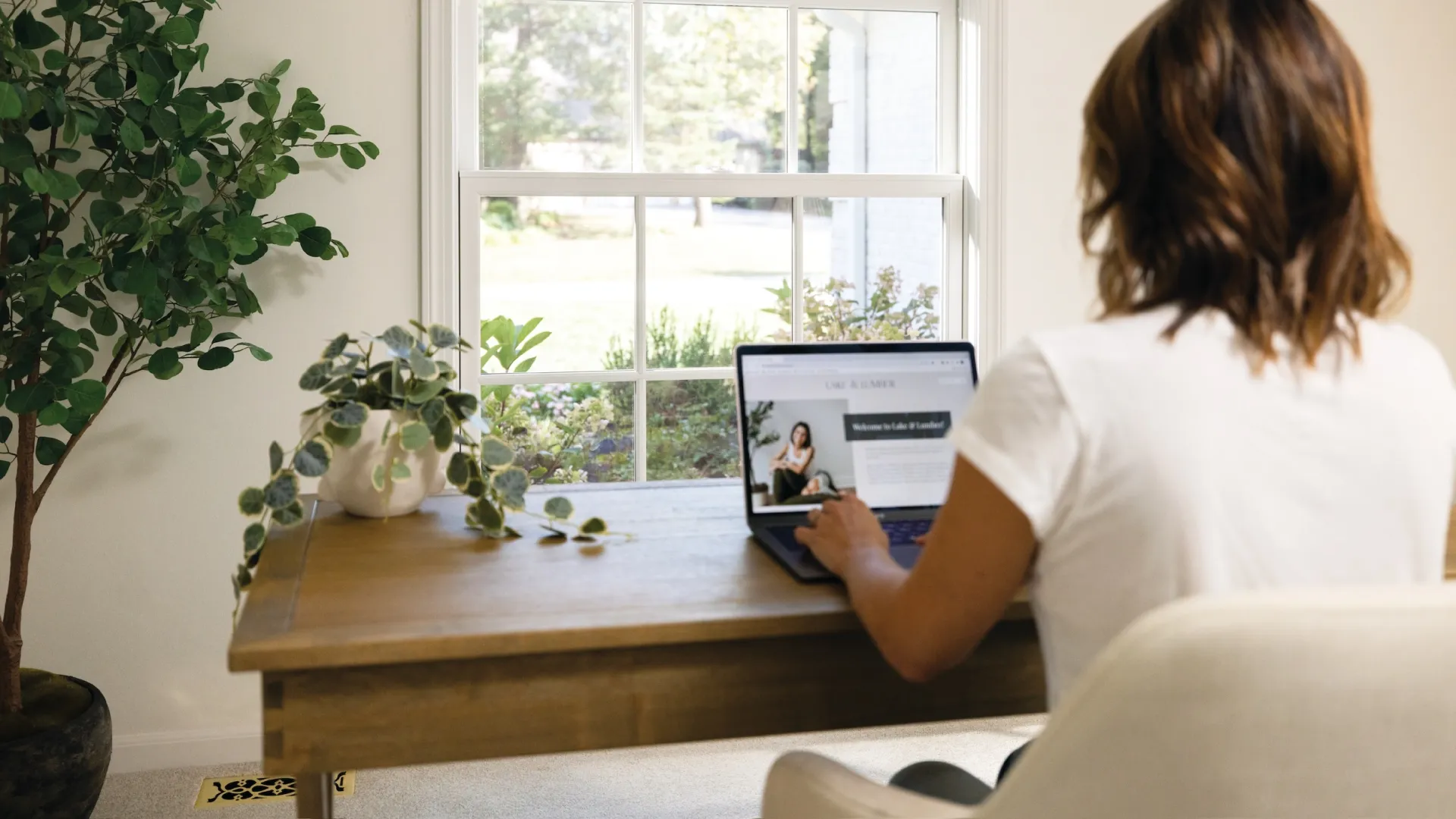 Person working on a laptop at a wooden desk near a large window with plants and natural light.