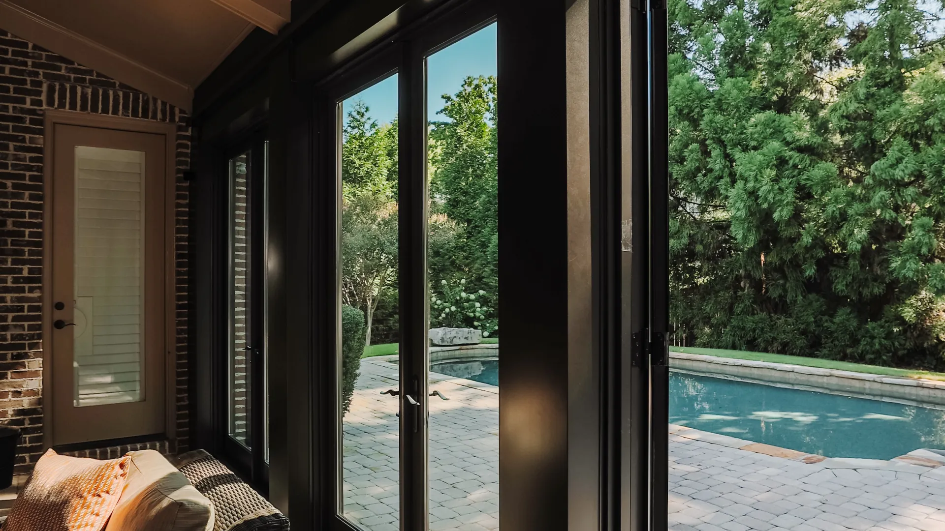 Cozy patio with black wicker furniture next to large glass doors opening to a pool and lush backyard.