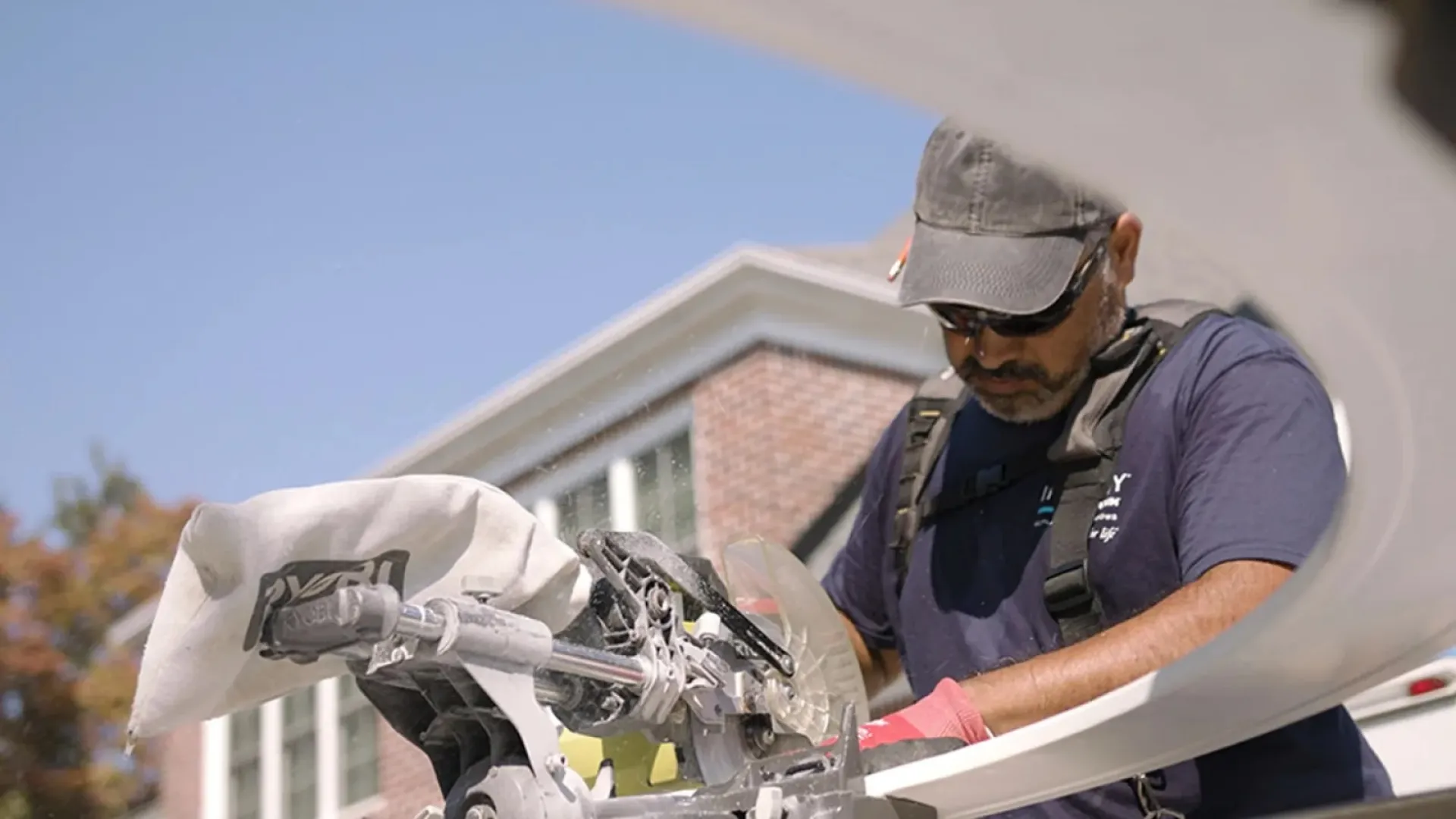 Skilled worker using a miter saw to cut wood outdoors near a brick house on a sunny day