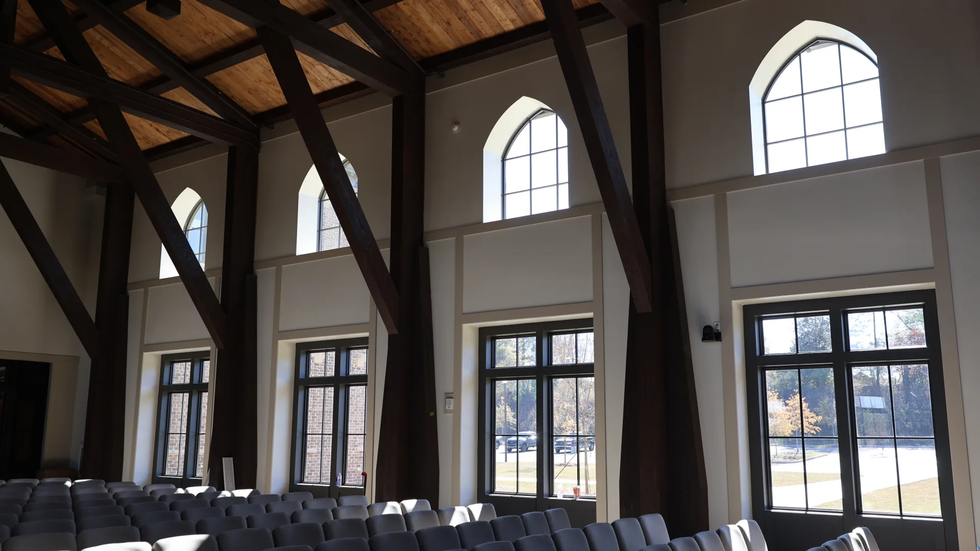 Interior of a spacious hall with large windows and wooden beams, featuring rows of gray chairs.