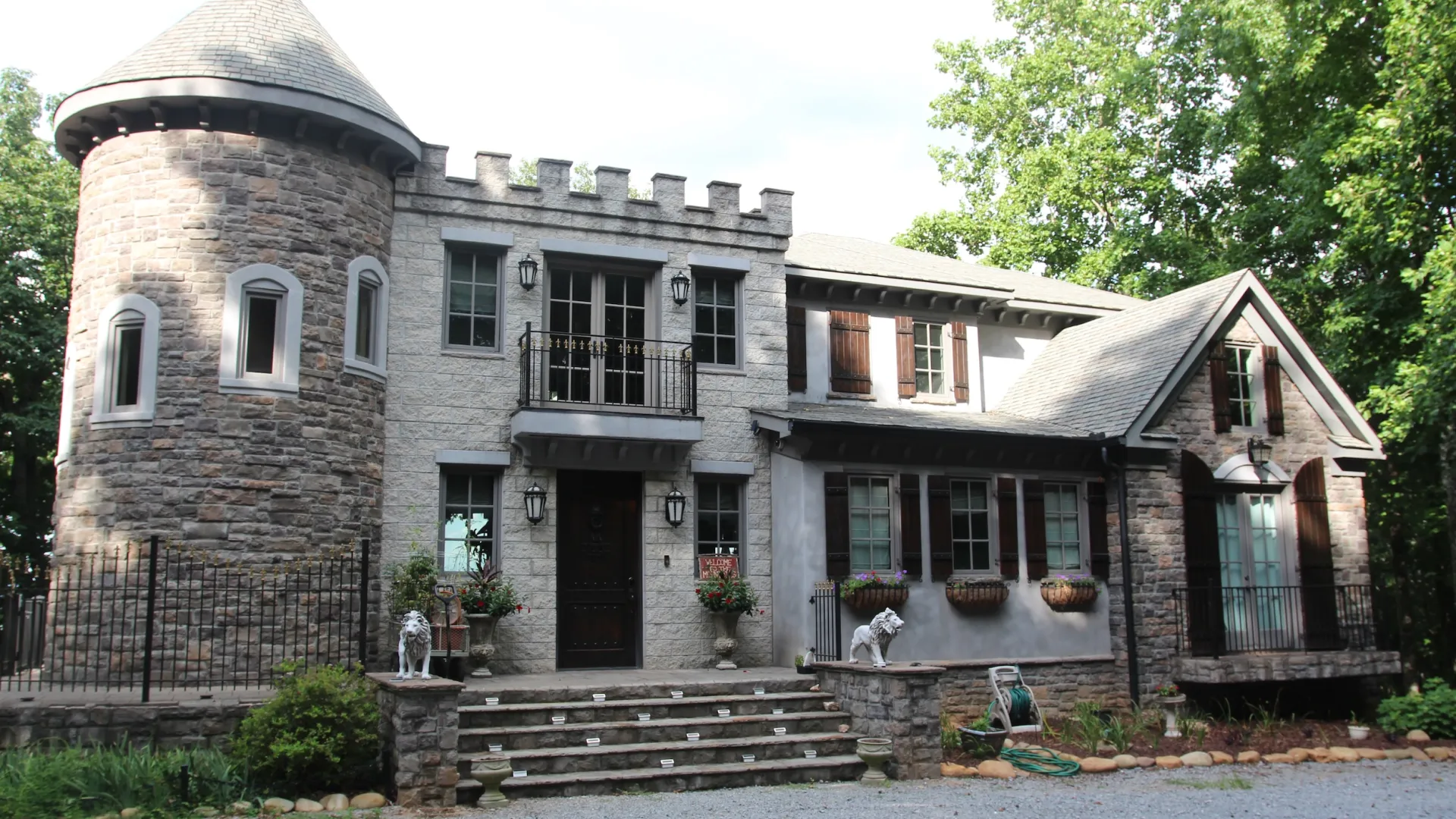 Stone castle-style house with turret, balcony, wooden shutters, and garden surrounded by trees and gravel driveway.