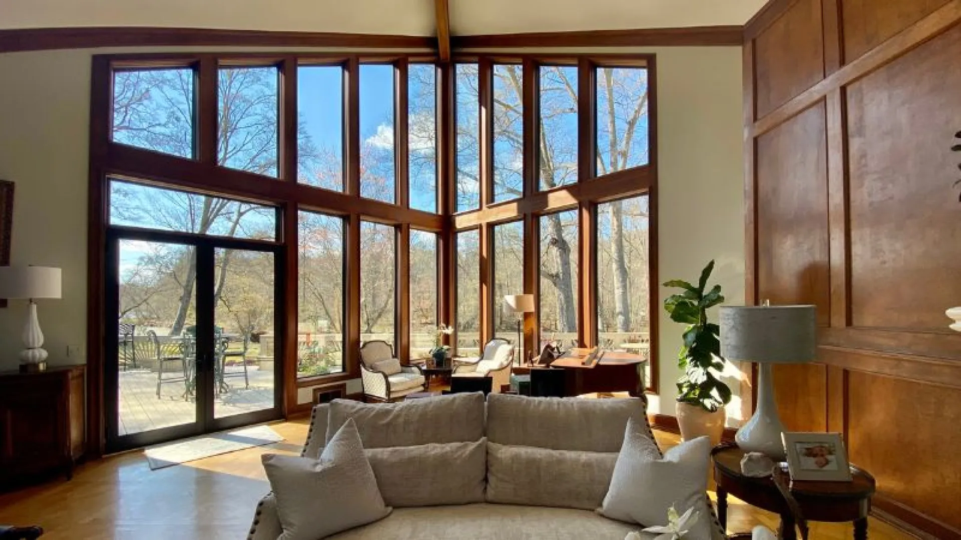 Bright living room with large floor-to-ceiling windows, cozy sofa, wooden accents, and outdoor trees view.