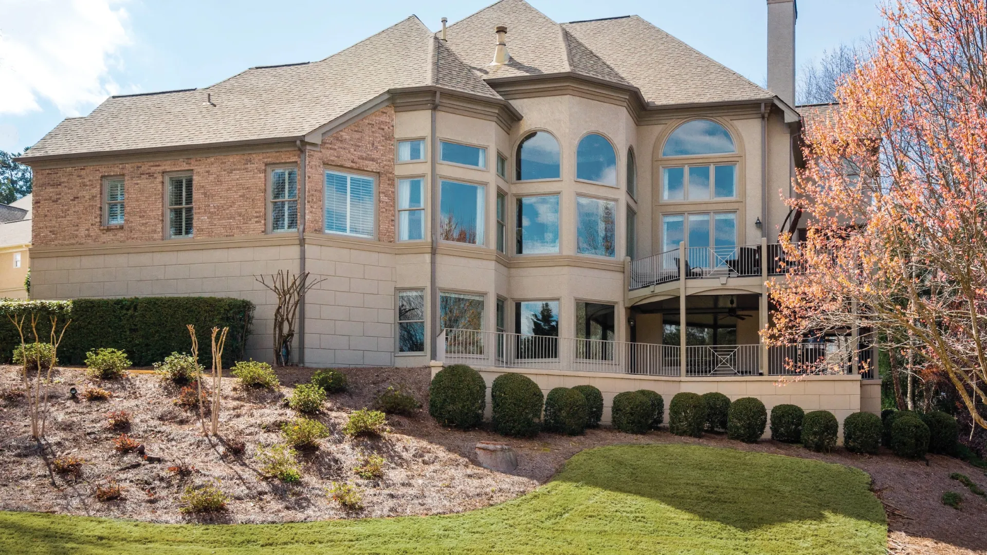 Large brick and stone two-story house with tall windows, a balcony, landscaped yard, and a blooming tree.