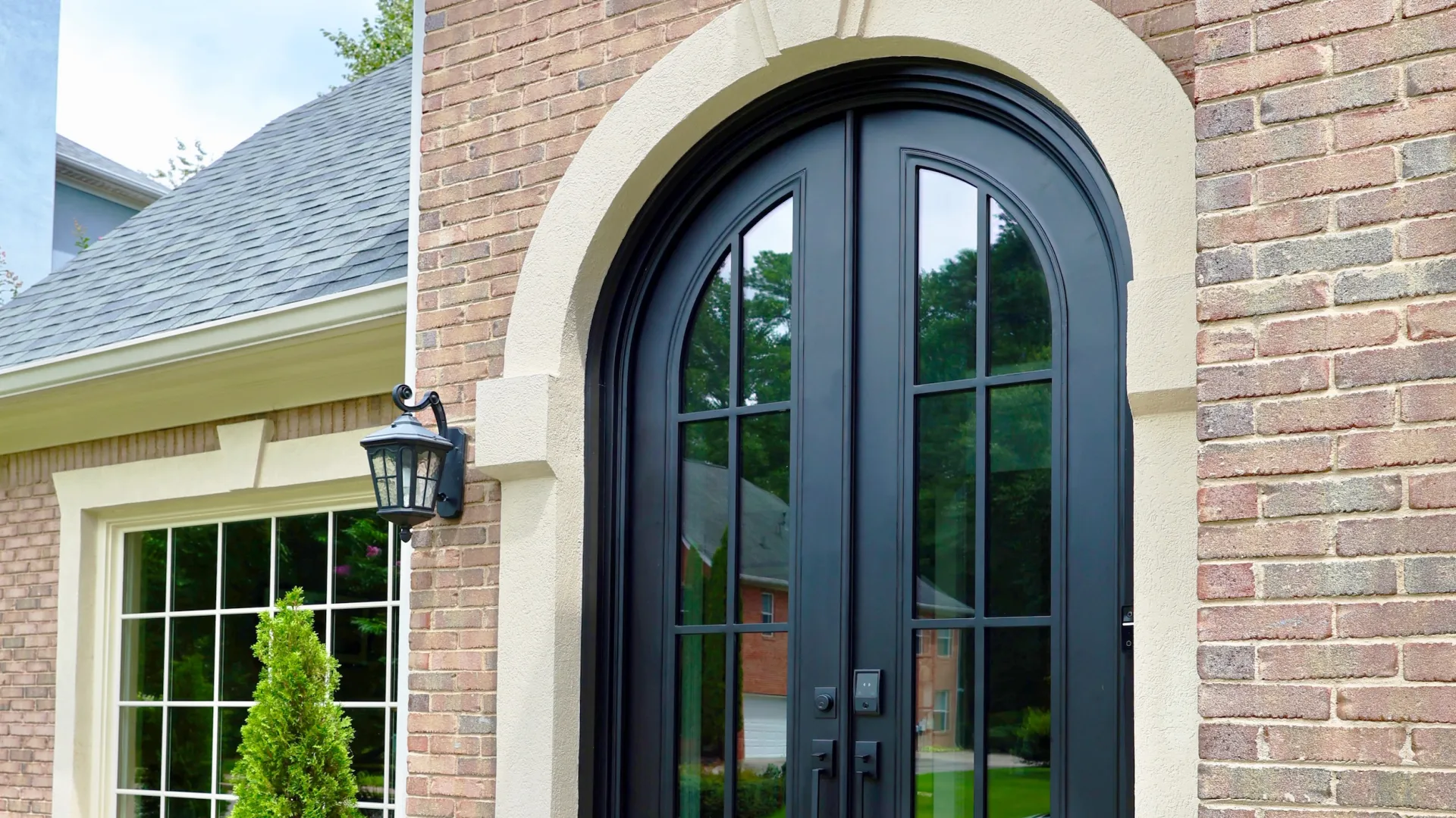 Black arched double doors with glass panels on a brick house entrance surrounded by greenery and flowers.