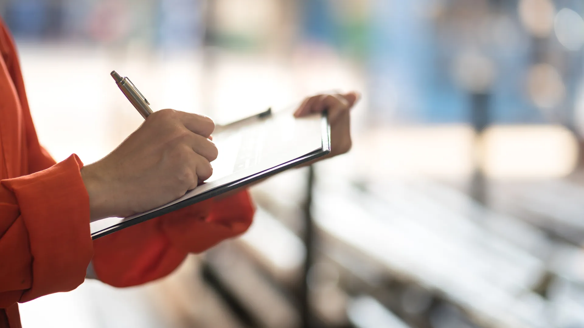 Person in orange sleeve writing on clipboard with blurred background of benches and sunlight.
