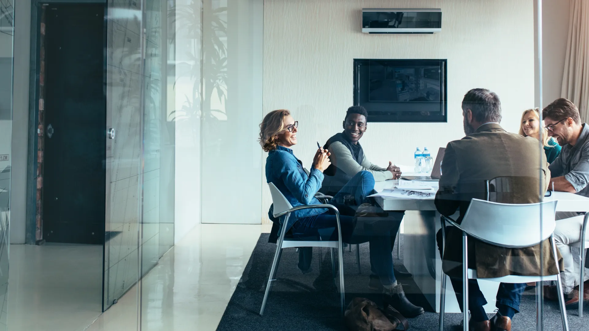 Five diverse professionals engaged in a meeting around a white table in a modern glass office.