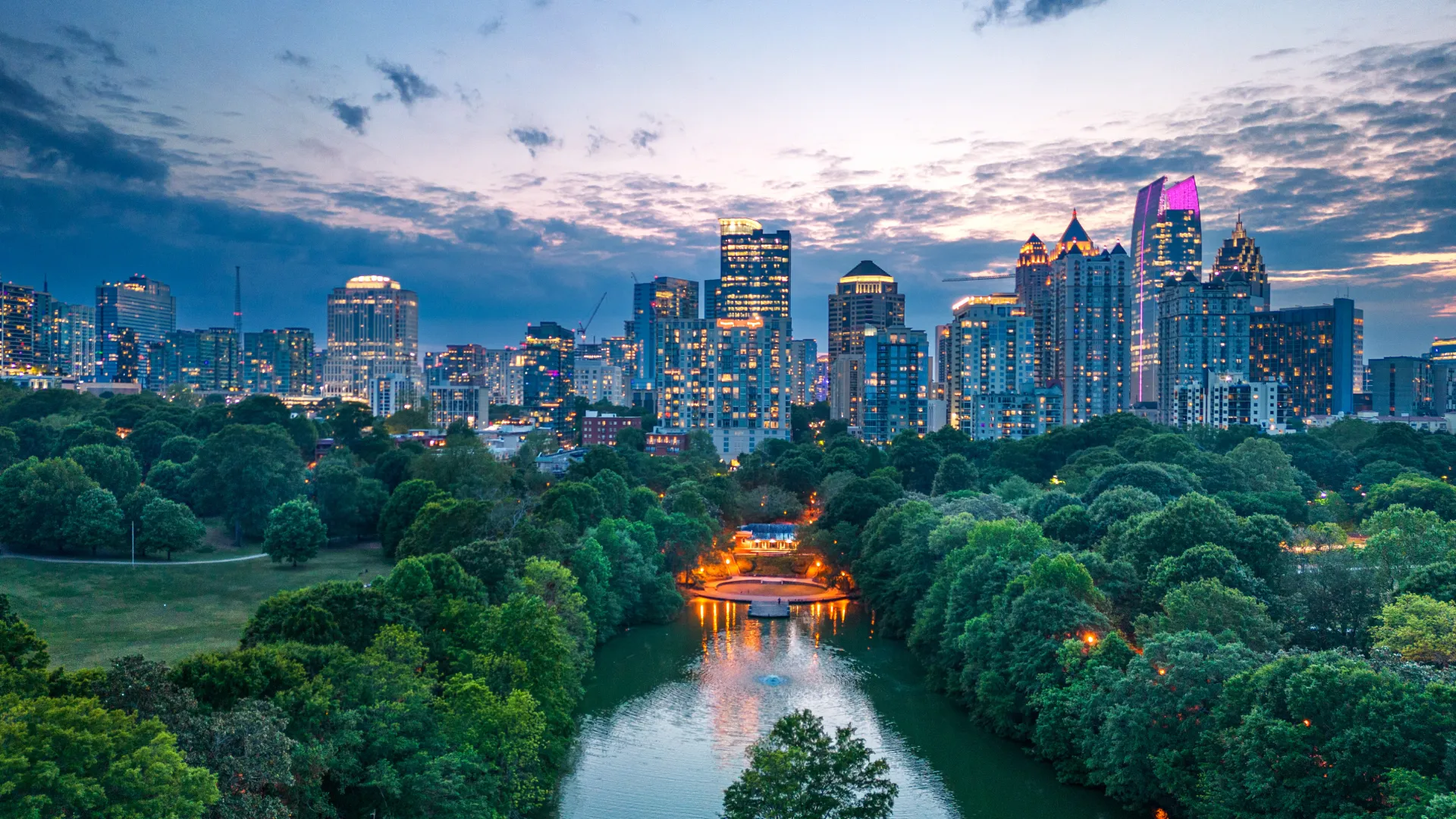 City skyline at dusk with illuminated skyscrapers and a river surrounded by lush green trees in foreground.