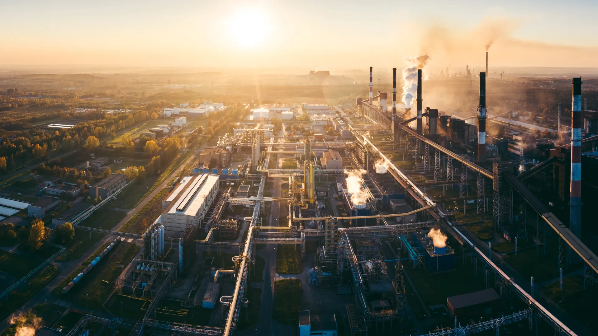 Aerial view of an industrial refinery complex with chimneys emitting smoke at sunset under a clear sky.