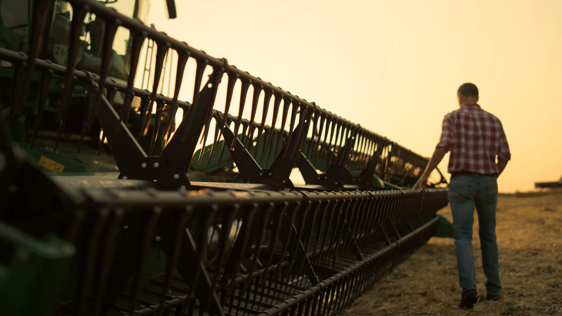 Farmer in plaid shirt standing next to a large combine harvester's cutting header at sunset in a field.