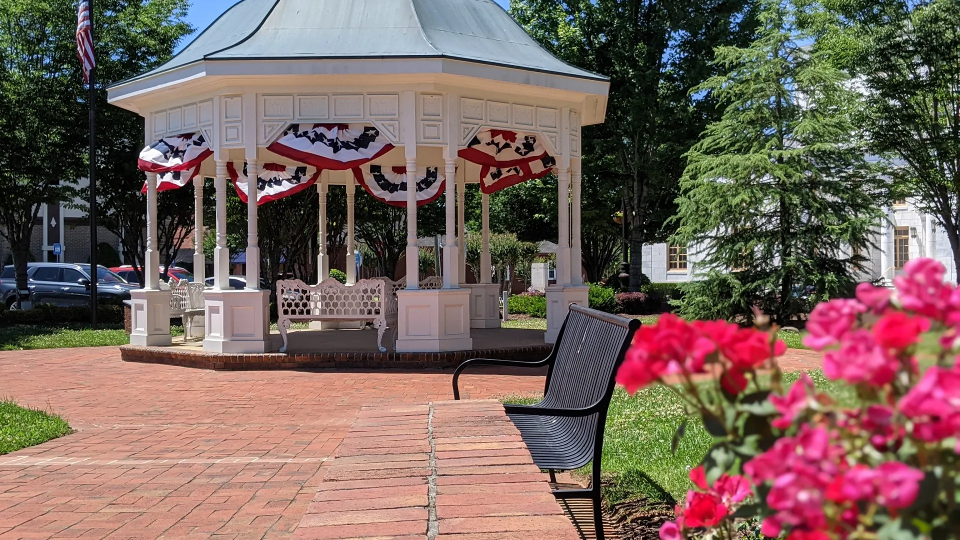 White gazebo decorated with patriotic bunting in a park with a bench and red flowers in foreground.