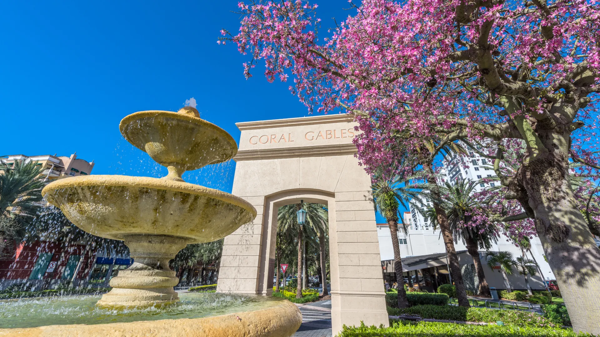 Fountain and archway with Coral Gables inscription surrounded by flowering trees and palm trees under blue sky