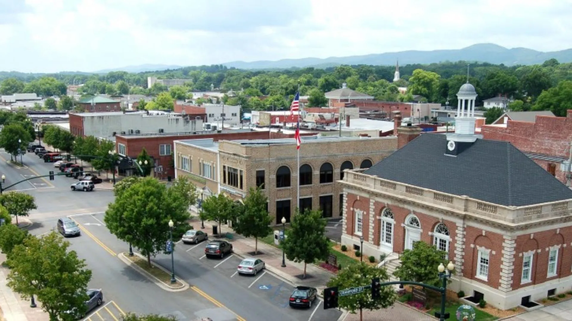 Small town downtown with historic brick buildings, American flag, trees, cars, and distant mountains under cloudy sky.