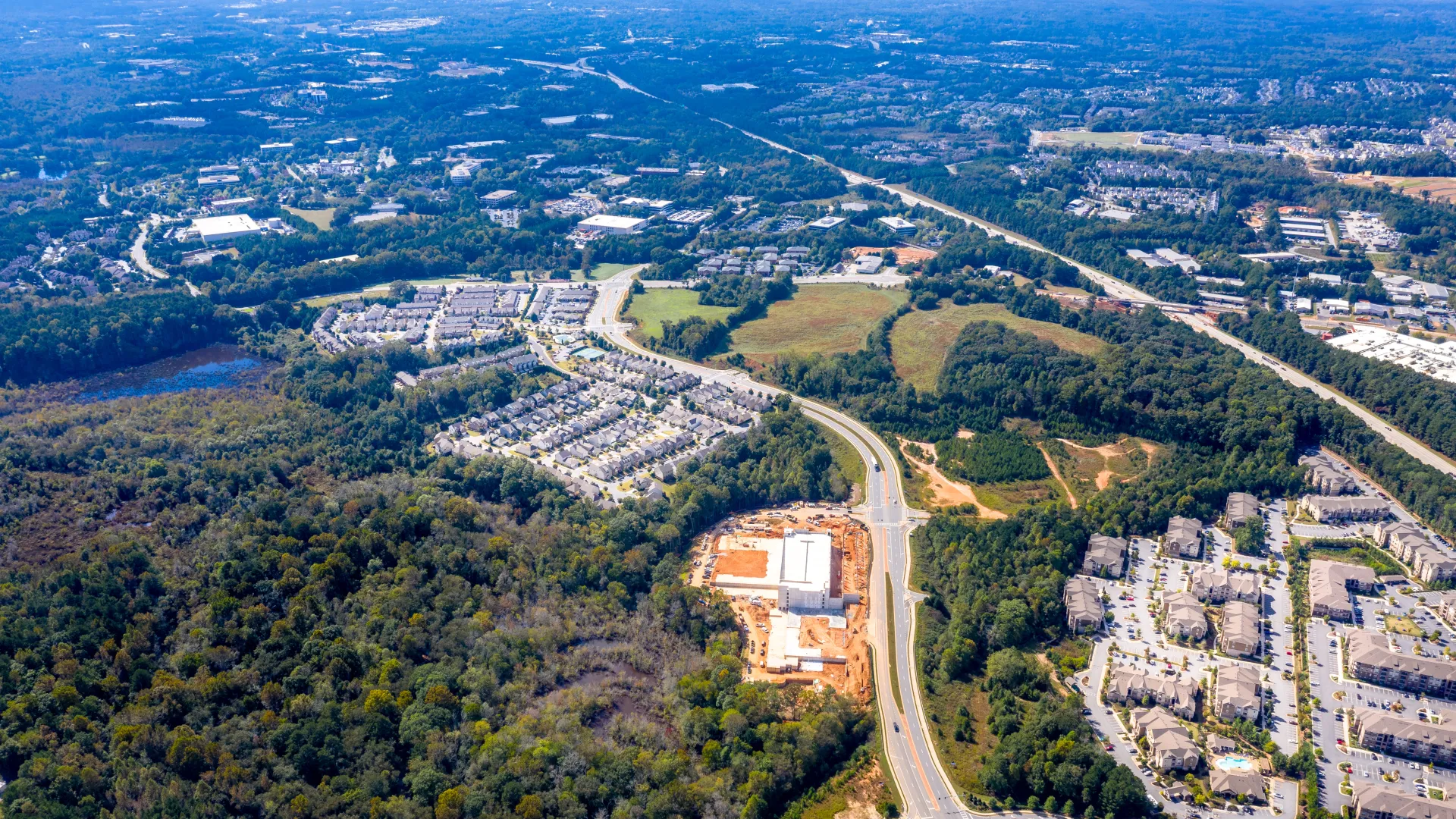 Aerial view of suburban neighborhood with roads, housing developments, forested areas, and clear blue sky.
