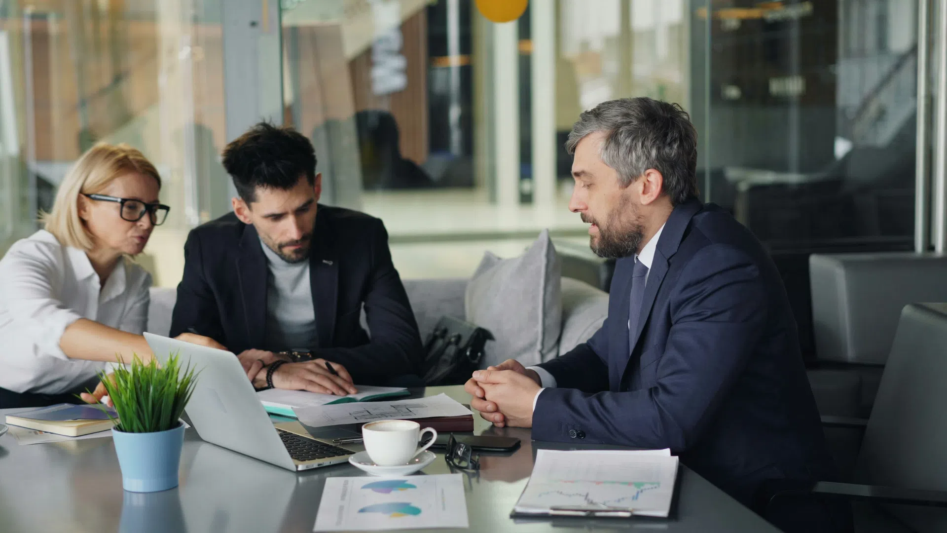 Three professionals in business attire discussing documents in an office meeting with laptop and charts on the table