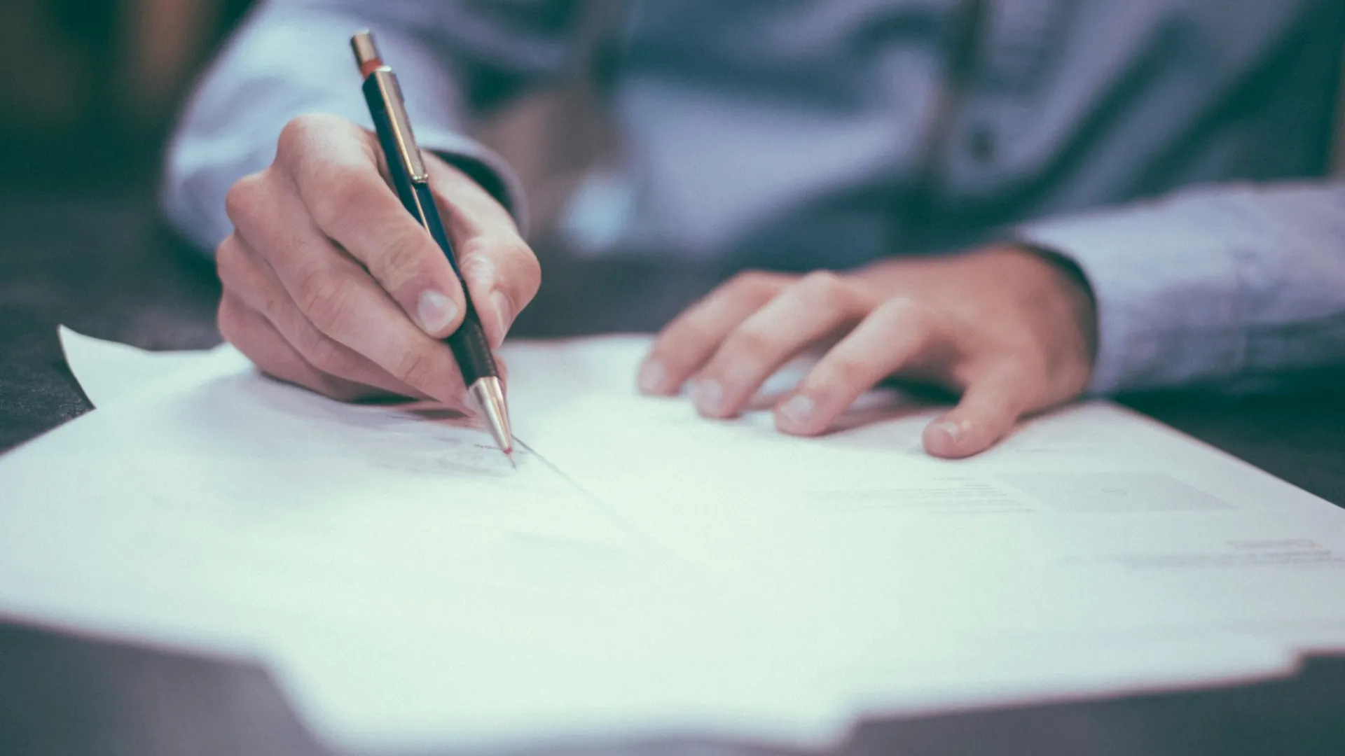 Person writing on documents with a pen, wearing a long-sleeve shirt, focus on hands and paper.