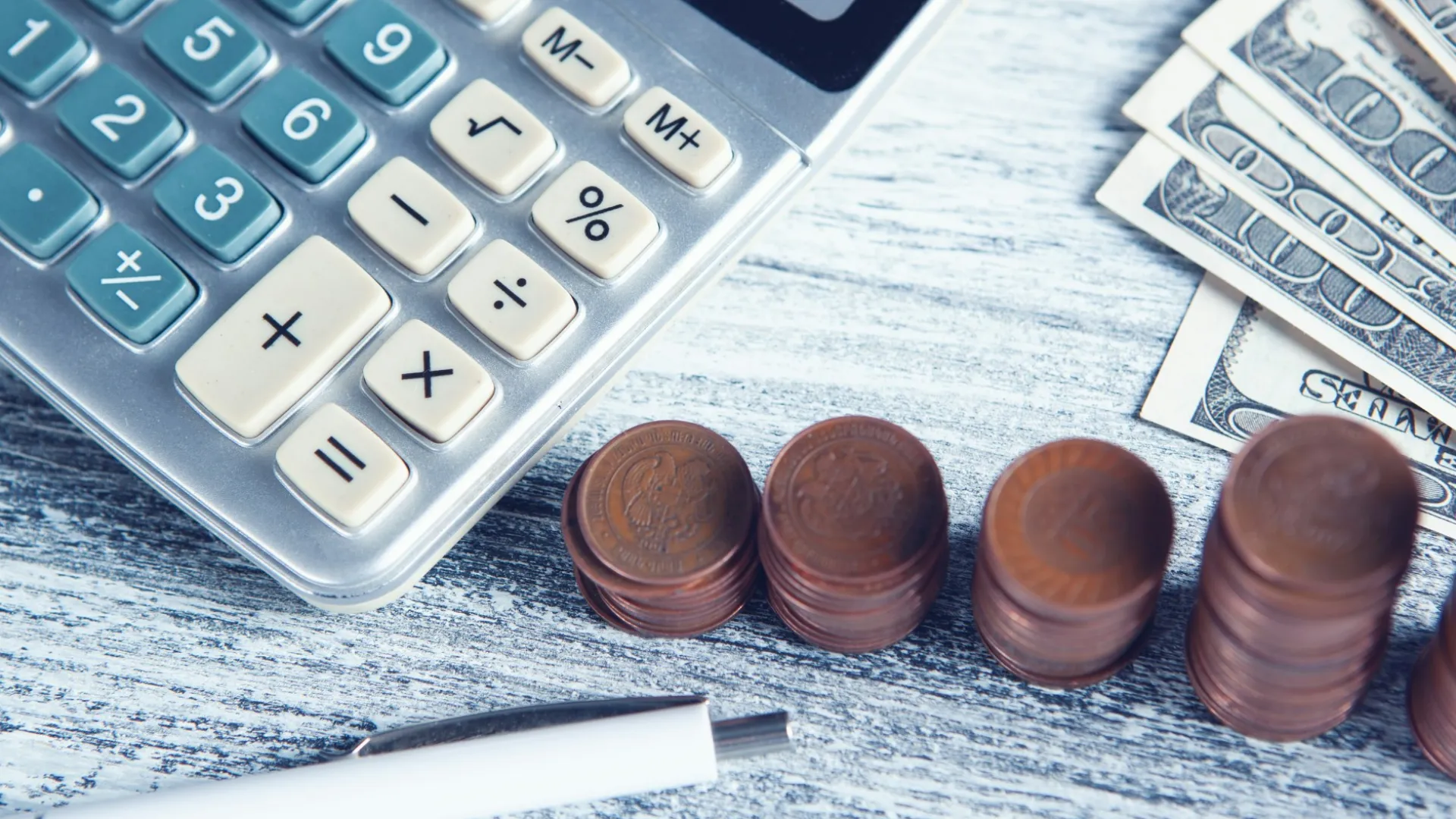Calculator, pen, stacked coins, and US dollar bills on a wooden table symbolizing finance and budgeting.