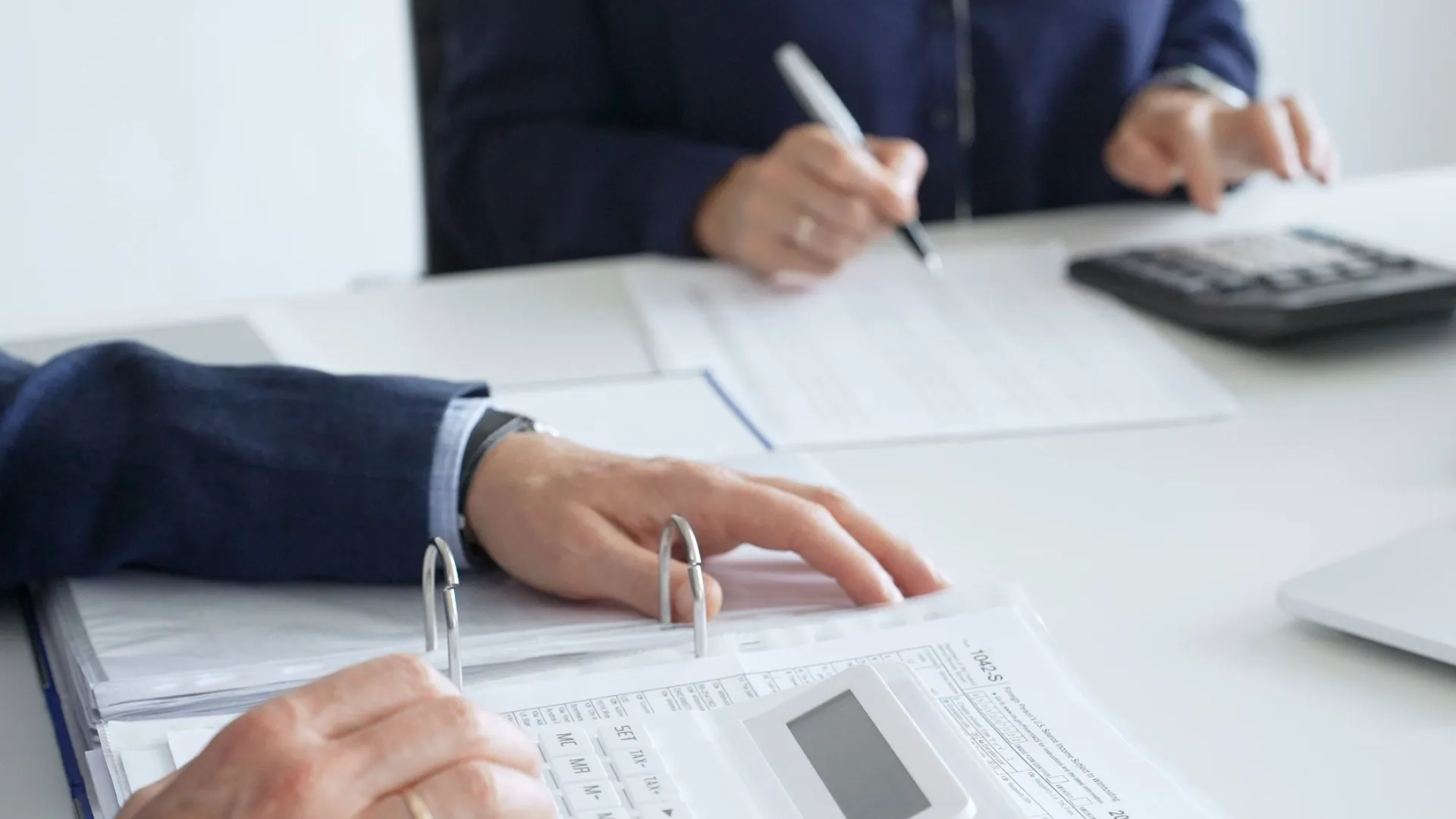 Two business professionals working with documents and calculator on office desk with laptop and files.