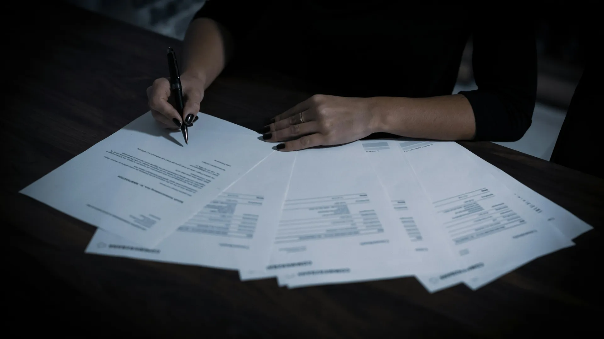 Person signing multiple documents spread out on a dark wooden desk in a low-lit setting.