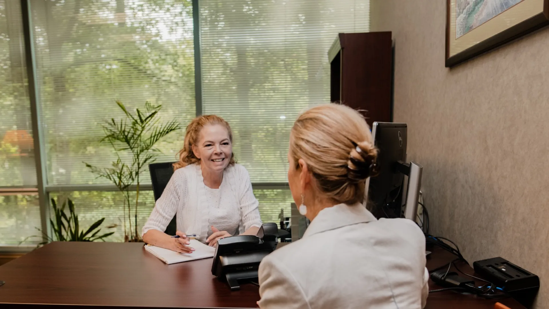 Two women in a professional office setting having a friendly consultation at a desk with computer and paperwork.