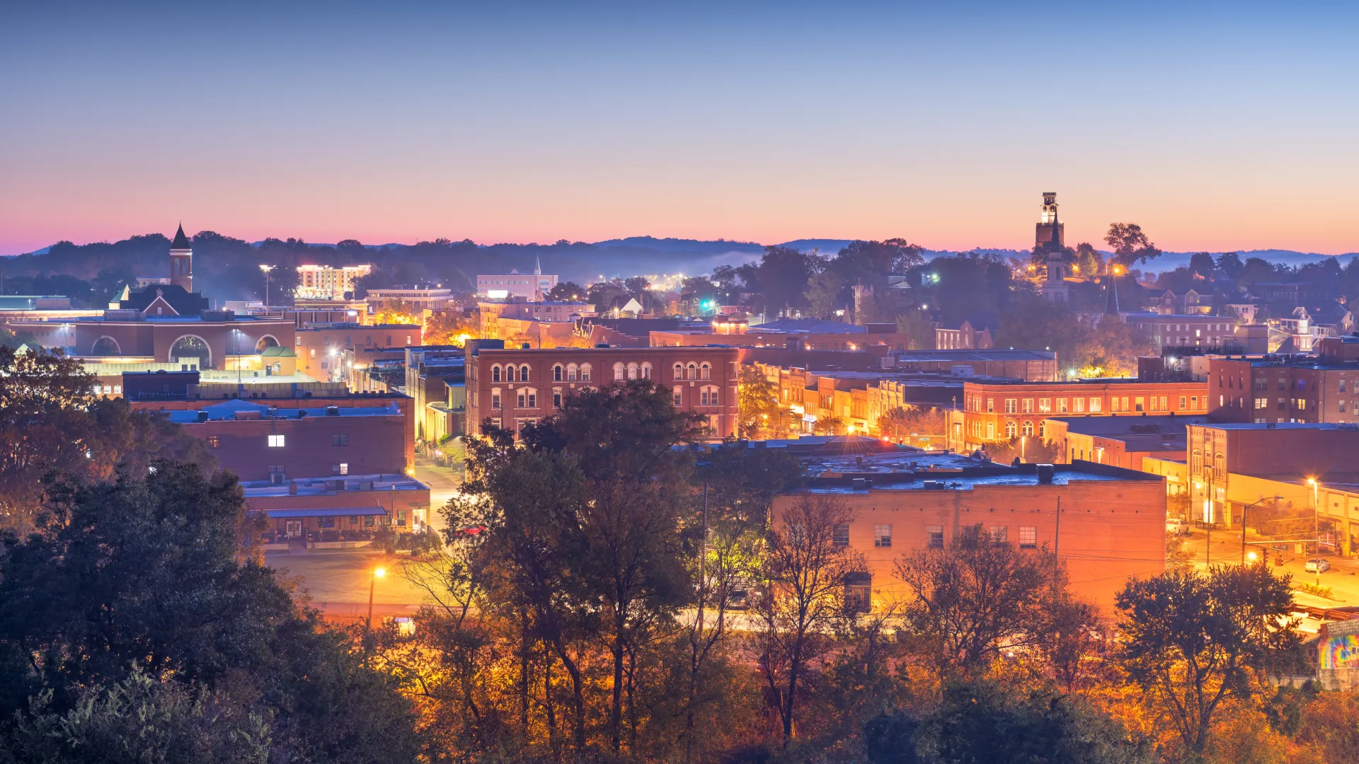 City skyline at dusk with illuminated buildings and a gradient sky transitioning from blue to orange.