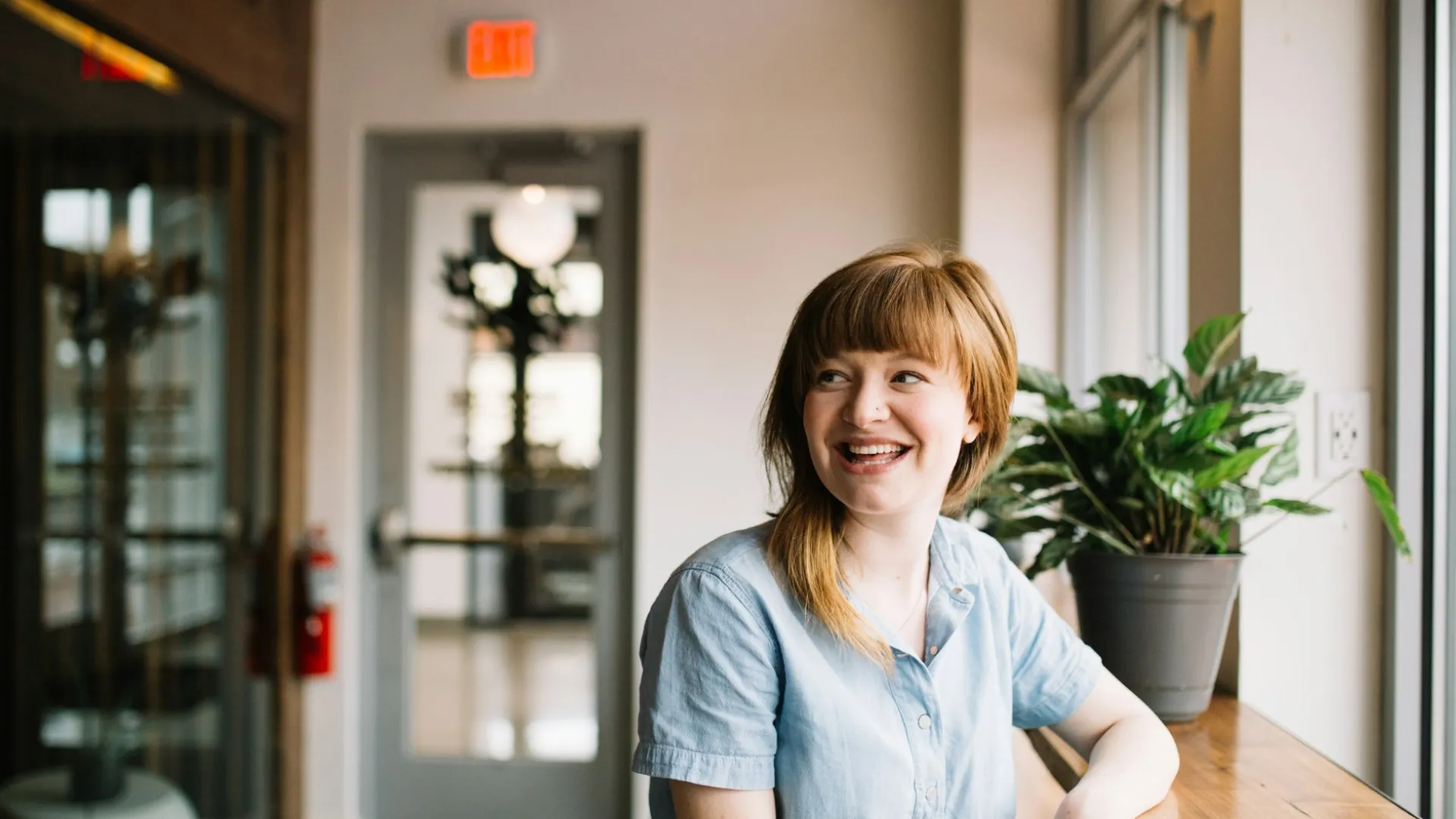 Smiling woman in light blue shirt sitting by a window with a plant and smartphone on the wooden counter