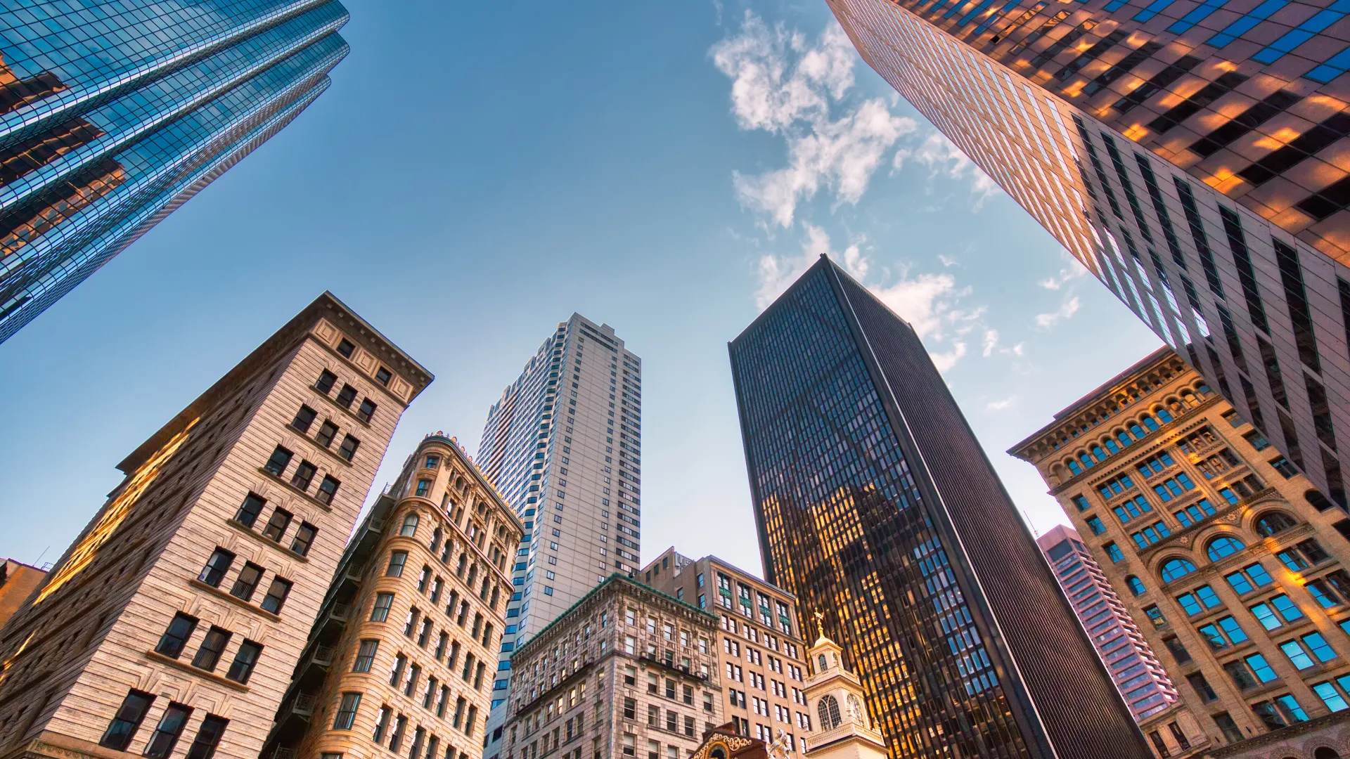 Low-angle view of modern and historic skyscrapers under a blue sky with scattered clouds in a cityscape.