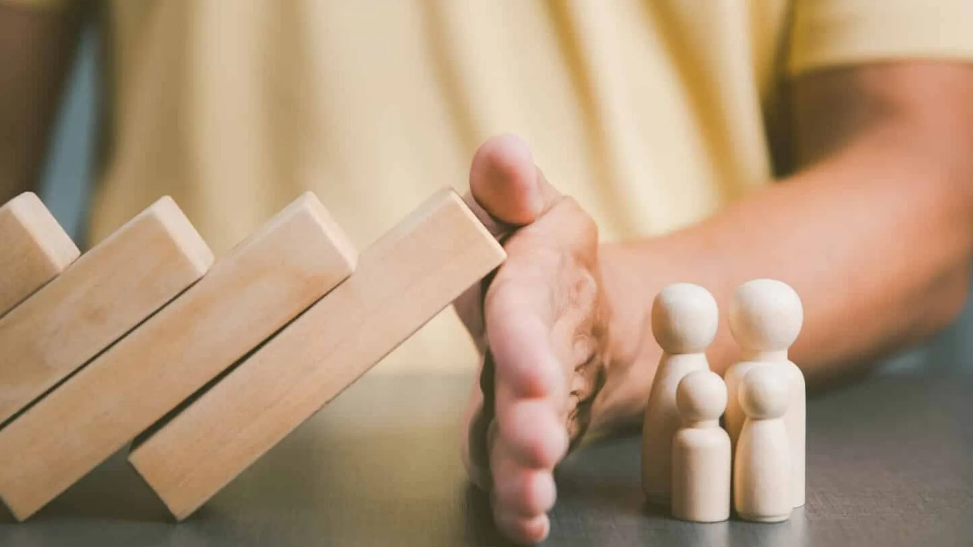 Hand stopping falling dominoes to protect small wooden family figures on a table, symbolizing safety and protection.