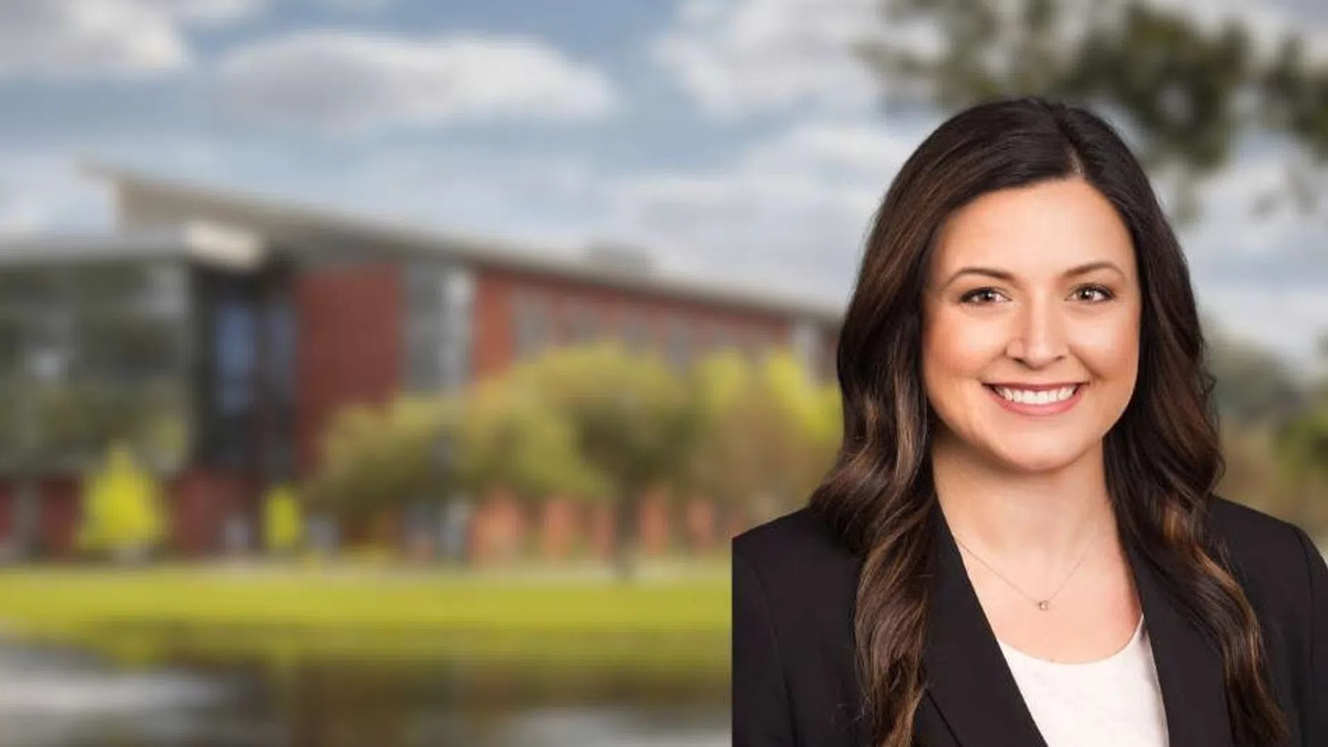 Professional woman smiling with a modern office building, trees, and water fountains in the background under a cloudy sky.
