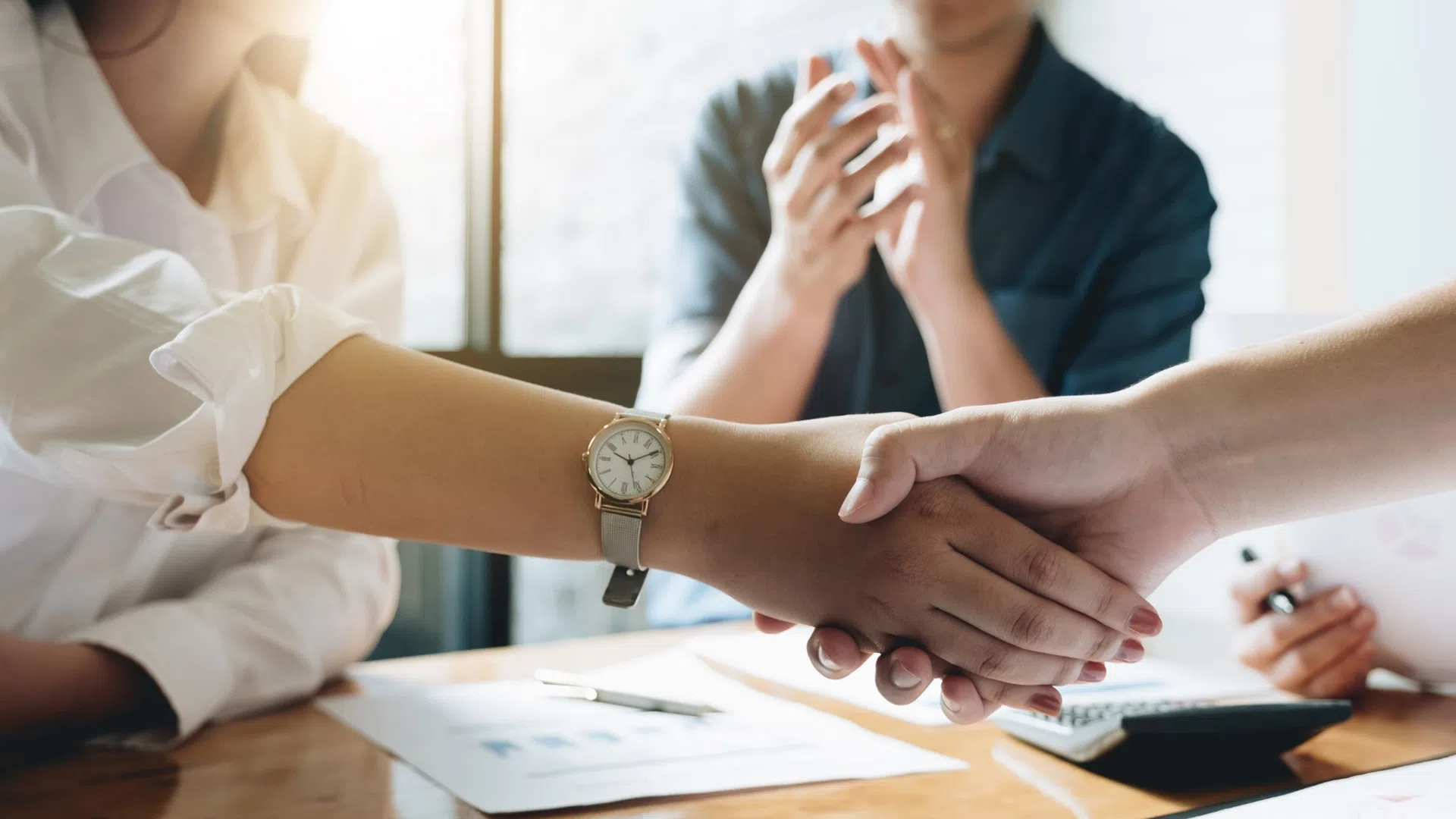 Two professionals shaking hands over business charts and laptops in a meeting room with natural light.