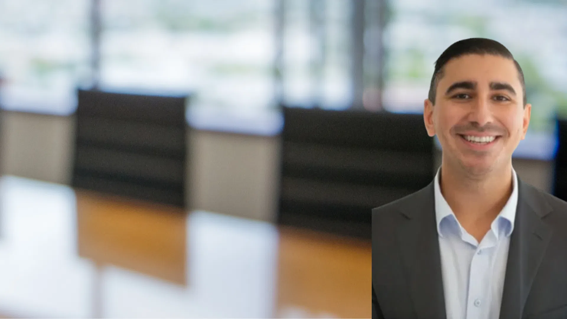 Smiling man in business suit with blurred office conference room and large windows in background.