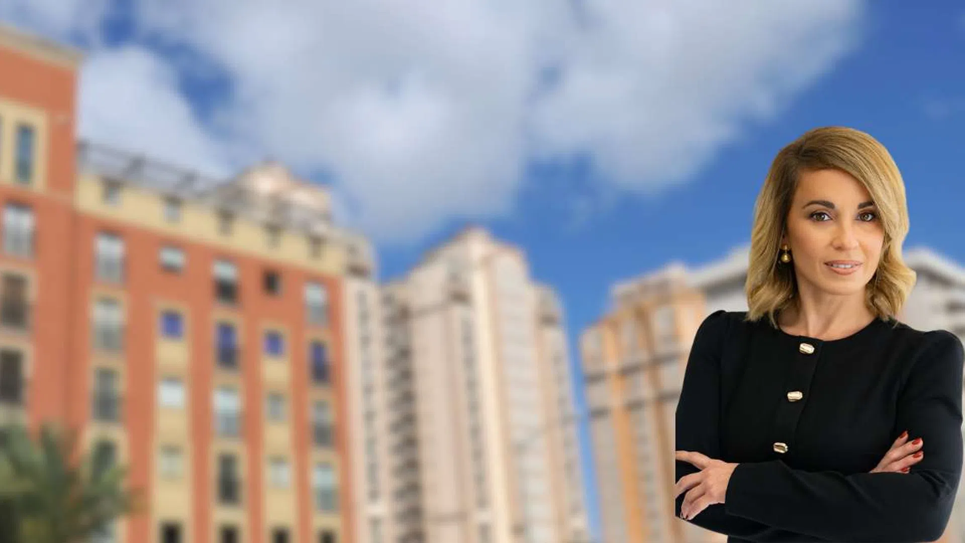 Confident businesswoman stands with folded arms in front of blurred city buildings under a blue sky.