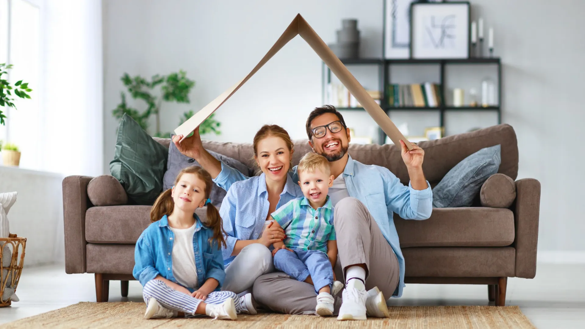 Happy family of four sitting on floor with parents holding roof frame symbolizing home and protection