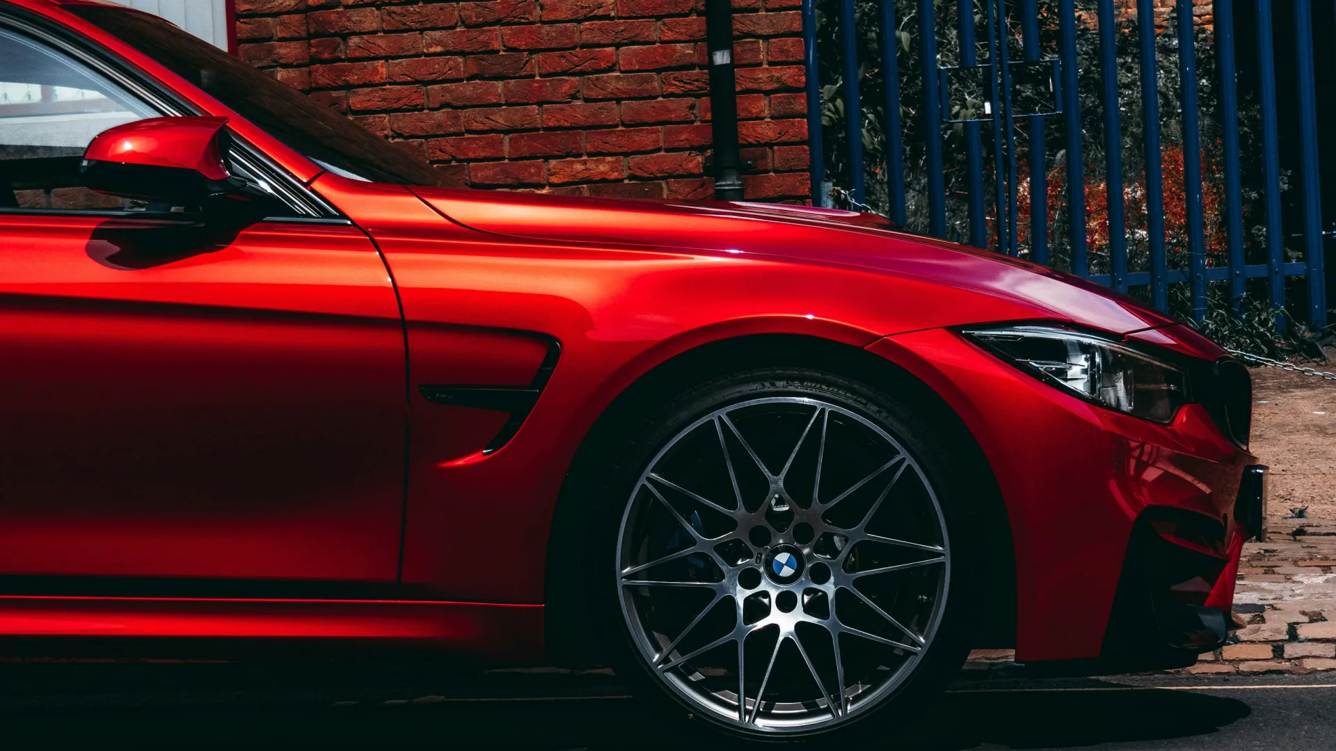 Sleek red BMW M4 parked against a brick wall with blue fencing, highlighting its sporty design.