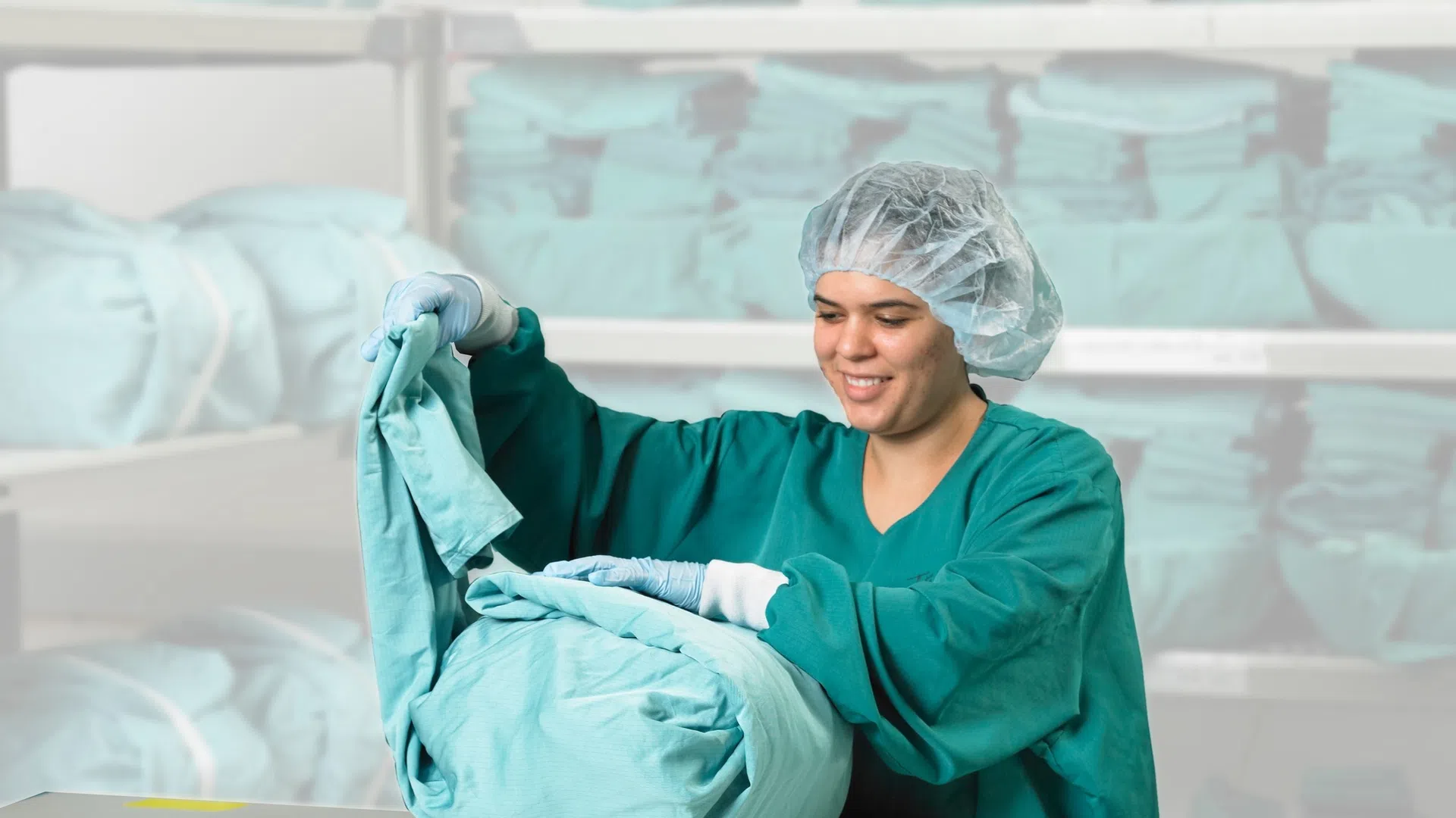 Healthcare worker in scrubs folding clean medical linens in a storage room with stacks of blue sheets.