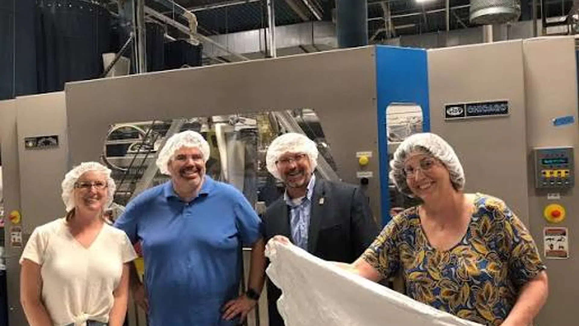 Four people wearing hairnets standing in front of industrial machinery, one holding a large sheet of dough or similar material