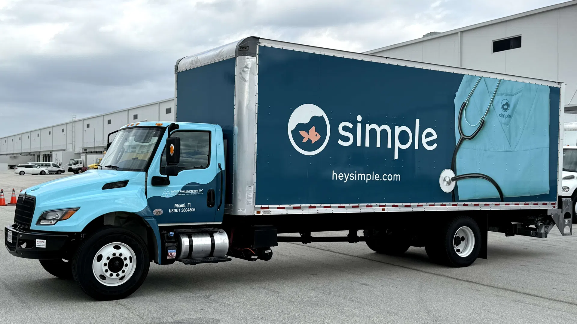 Light blue and dark blue Simple branded delivery truck parked outside a warehouse with stethoscope graphic on the side.