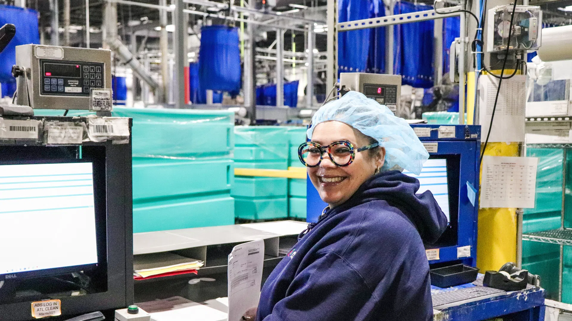 Smiling factory worker wearing hairnet and glasses works at computer station surrounded by industrial equipment.