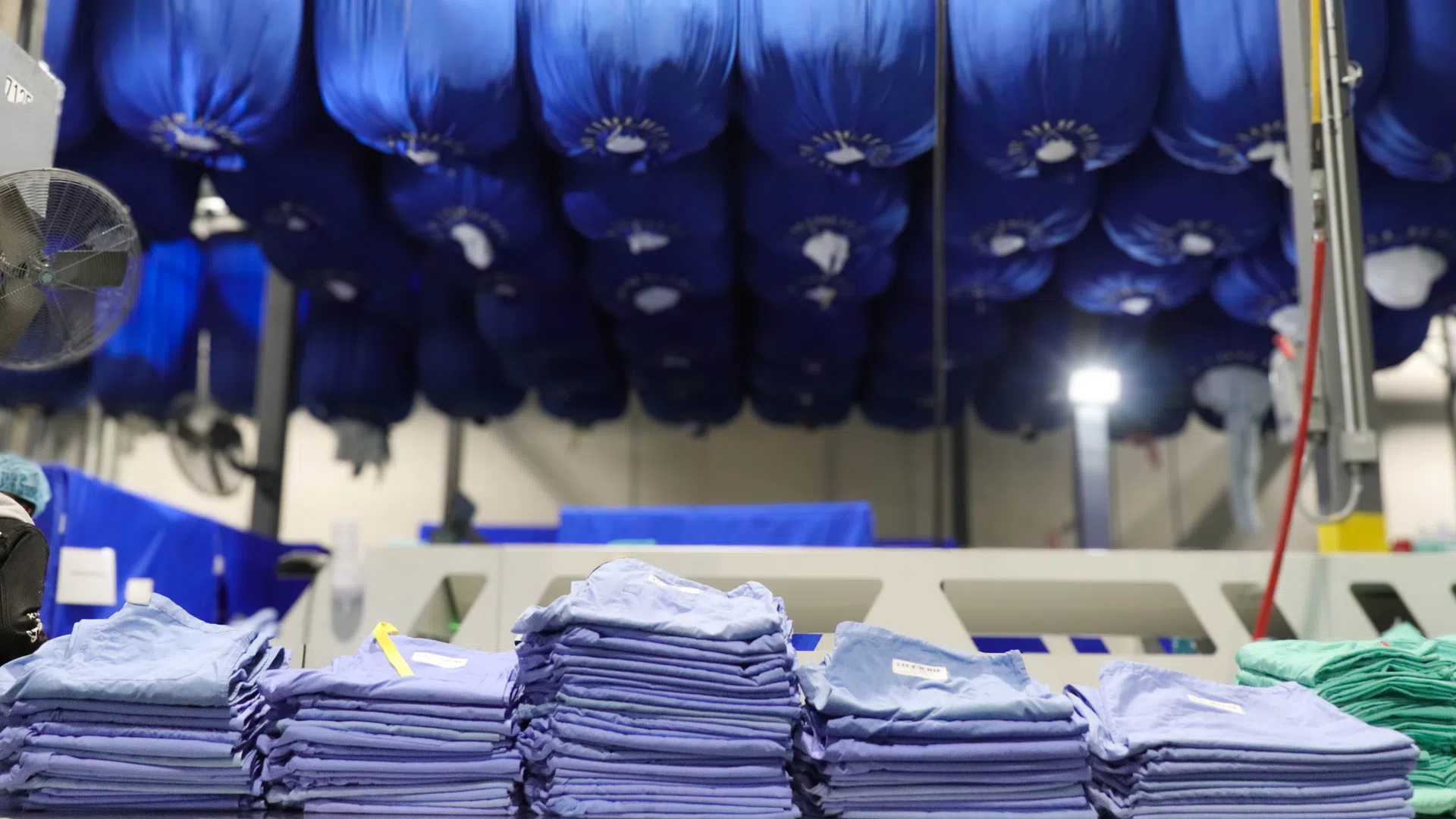 Stacks of folded blue and green garments on a table with blue garment bags hanging from ceiling in a laundry facility.