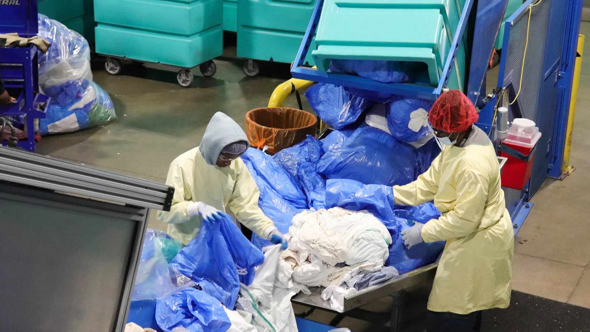 Workers in protective gear sorting blue and white laundry in an industrial laundry facility with large carts.