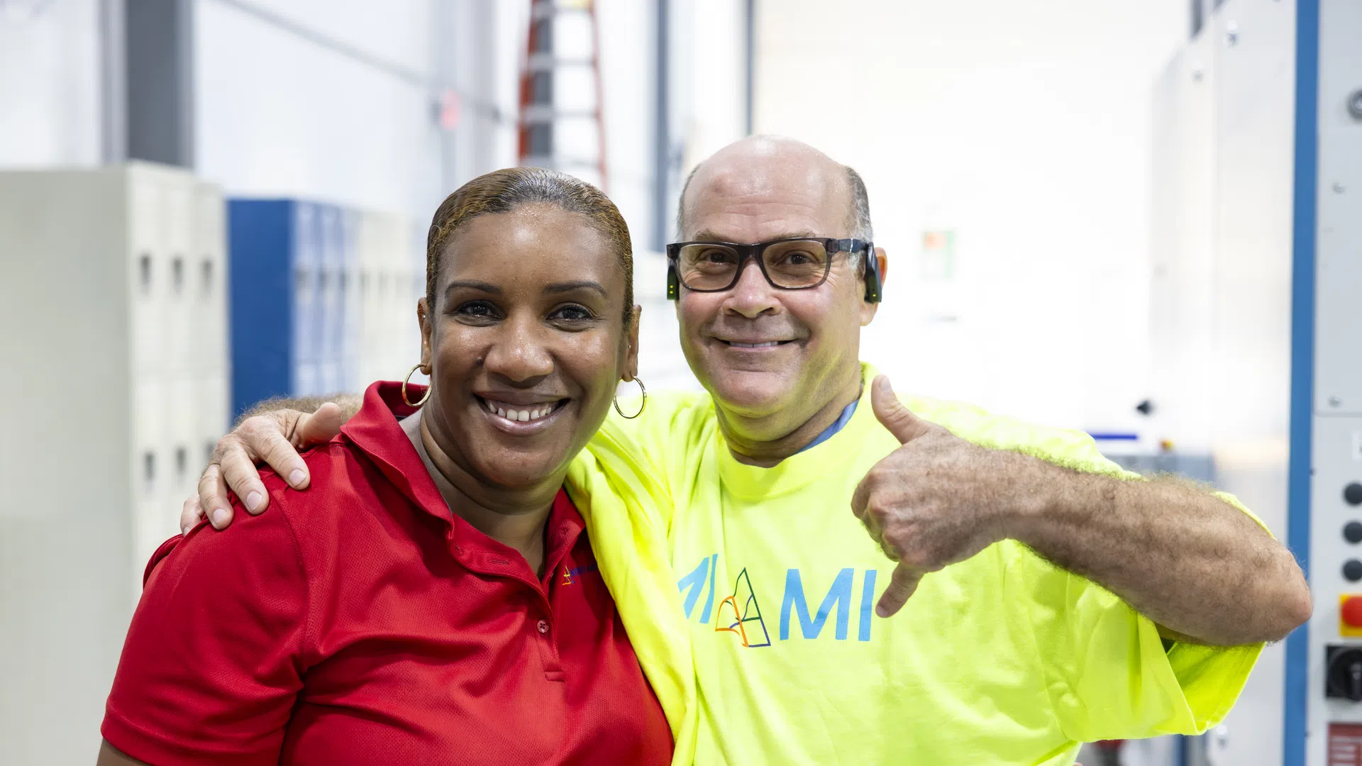 Two factory workers smiling together, one in red shirt and the other in yellow, showing a thumbs-up gesture indoors.