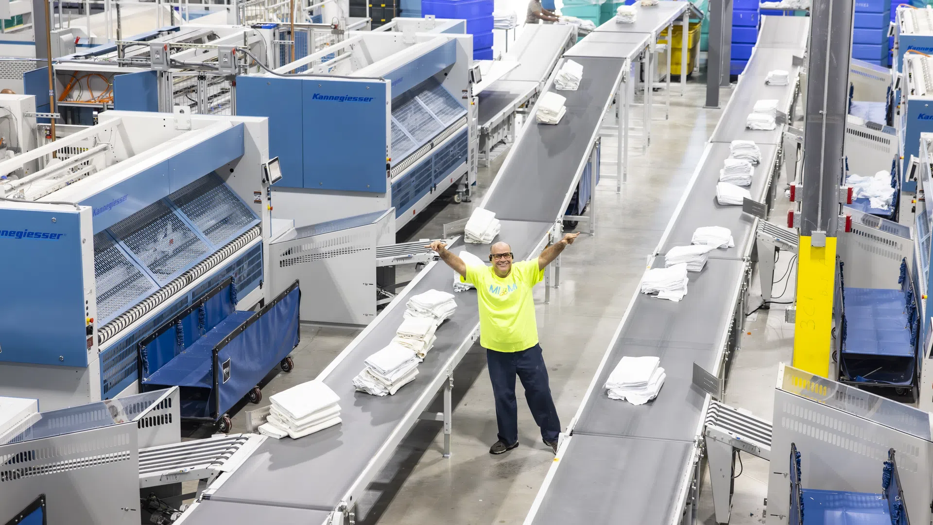 Industrial laundry facility with conveyor belts sorting folded white linens and a worker in a neon yellow shirt raising arms.