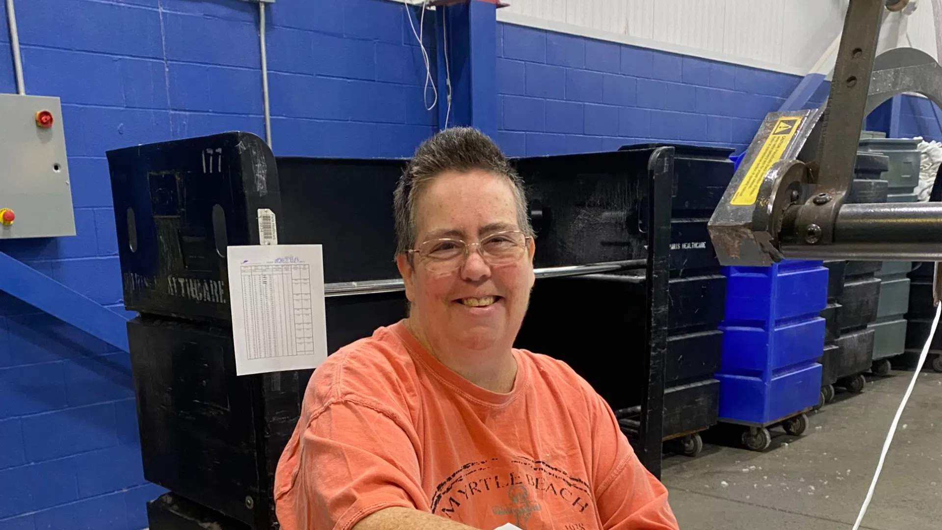 Smiling person folding white linens on a metal table in a blue-walled industrial laundry facility.