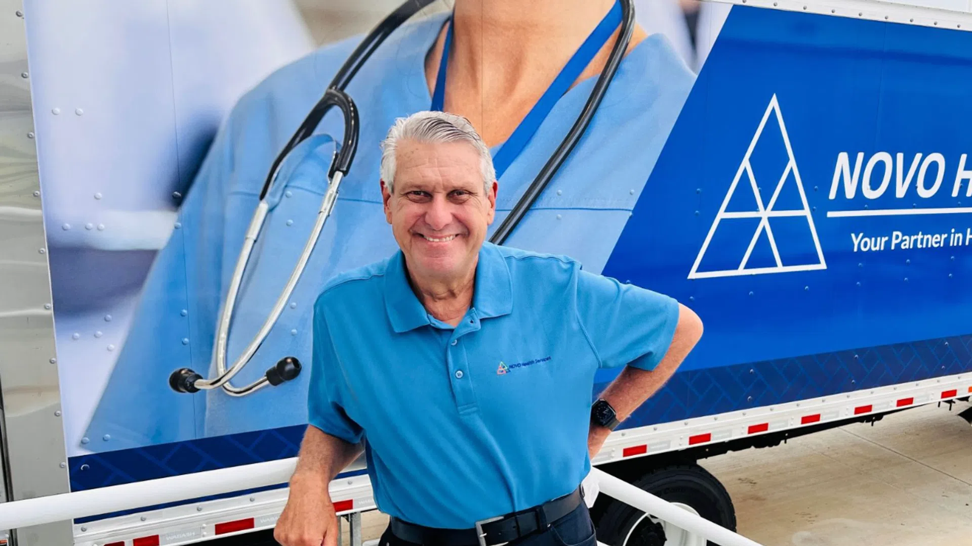 Smiling man in blue polo shirt standing in front of Novo Health Services truck with medical logo and stethoscope image.