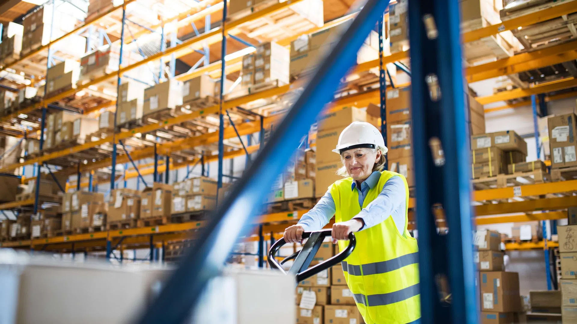 Warehouse worker in a safety vest and helmet operating a pallet jack among shelves of boxed inventory.