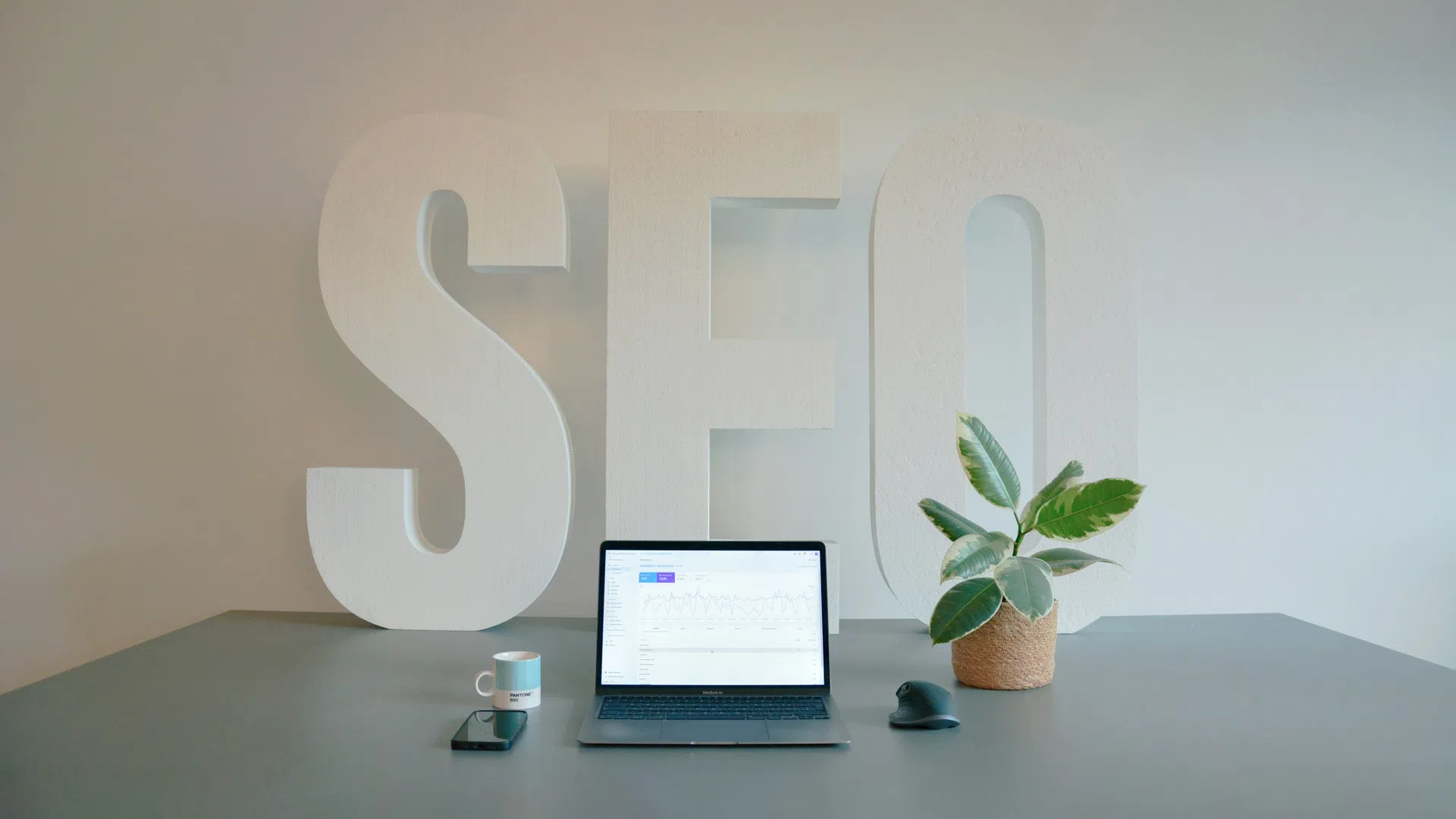 Workspace with laptop showing SEO data, smartphone, coffee mug, plant, and large white SEO letters in background.