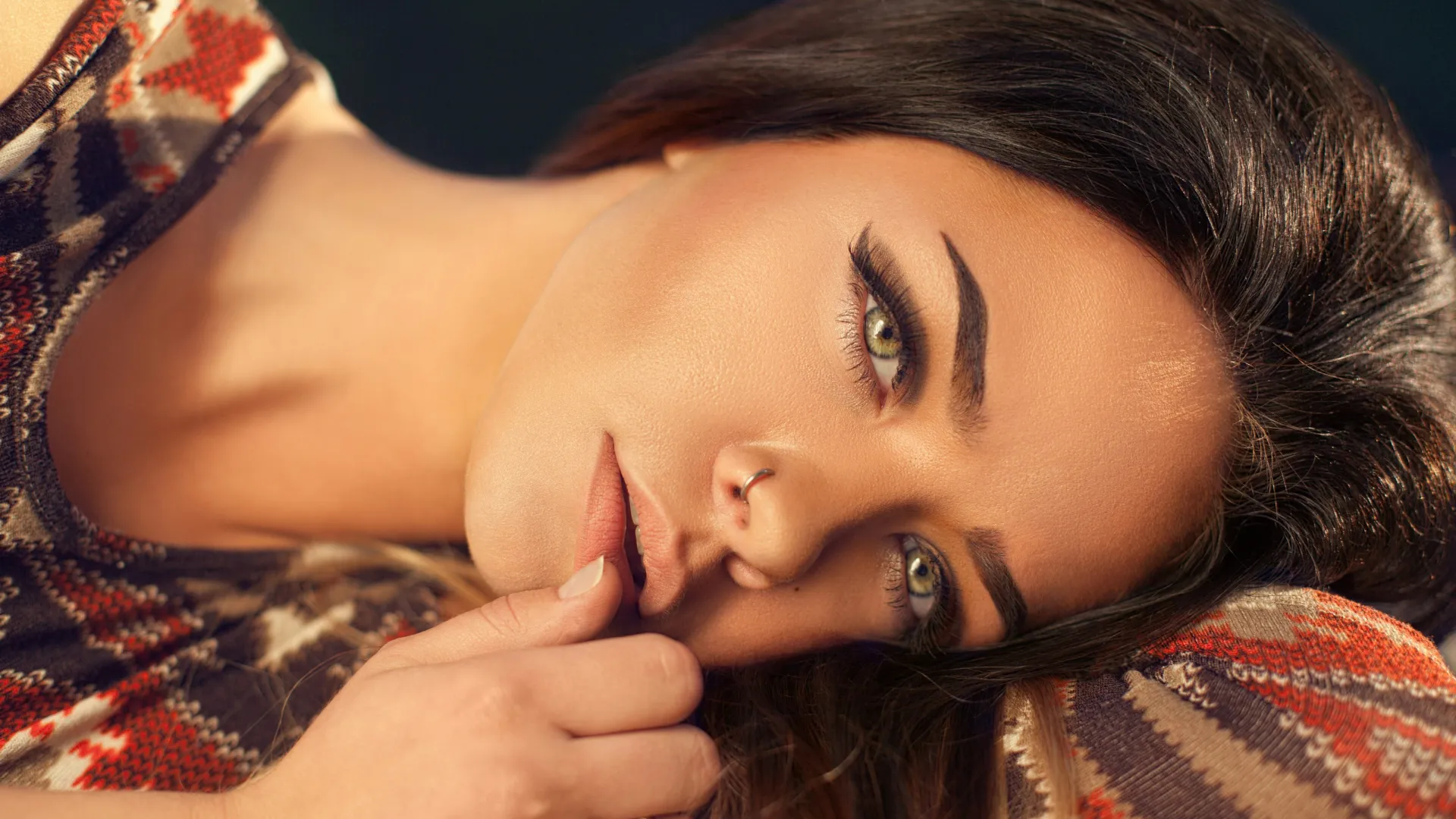 Close-up of a woman with green eyes and dark hair lying down with a patterned sweater and nose ring