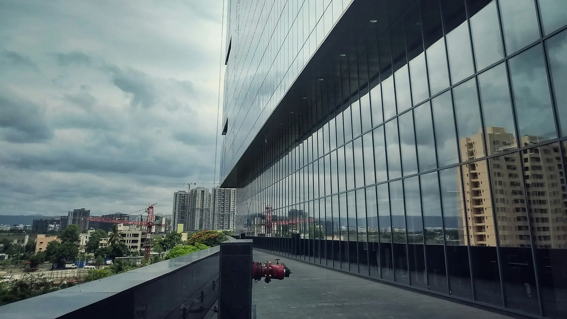 Modern glass building reflects nearby cityscape under a cloudy sky with urban buildings in the distance.