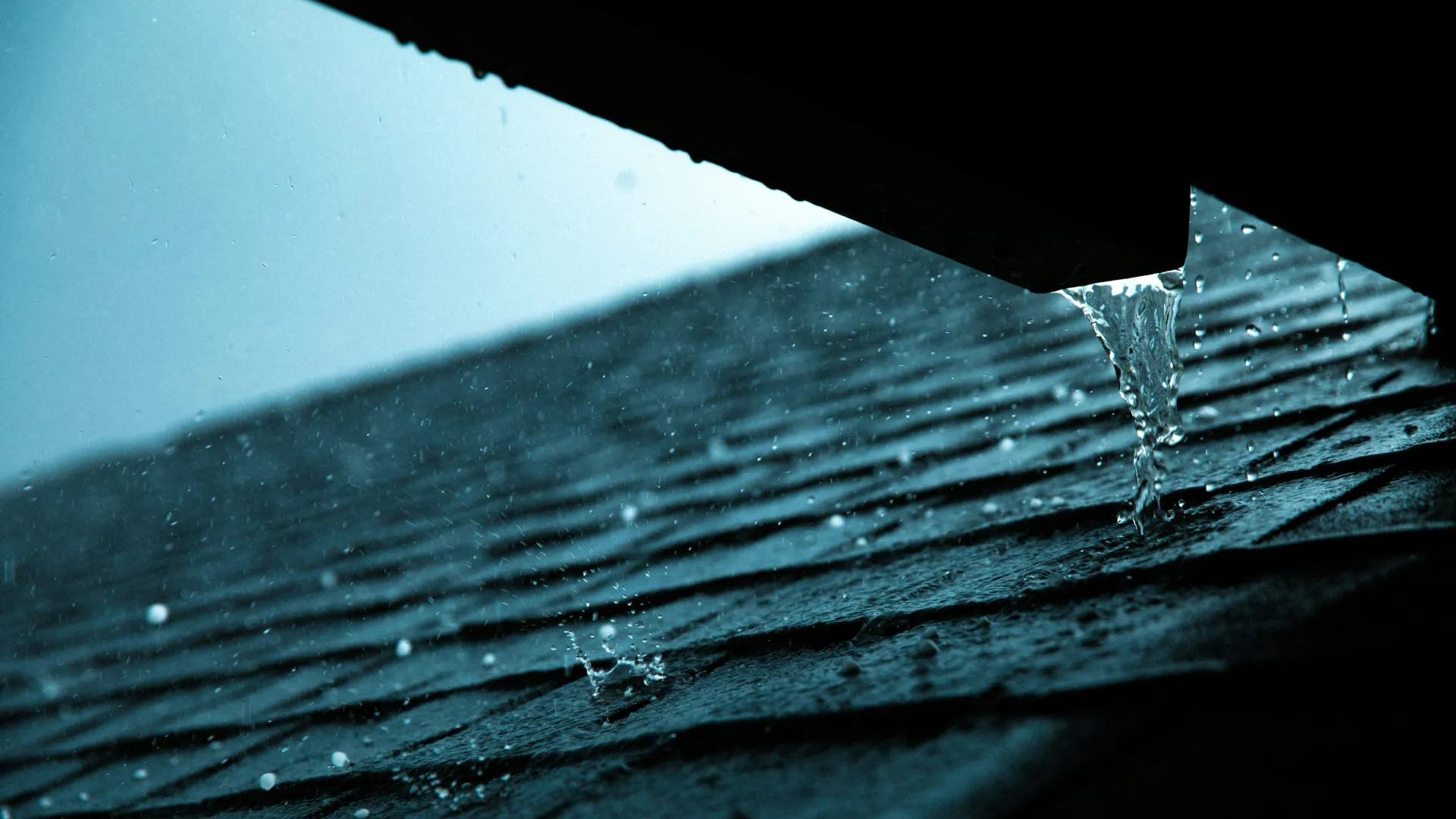 Water droplets falling from a roof edge against a rainy blue-gray sky with wet shingles below.